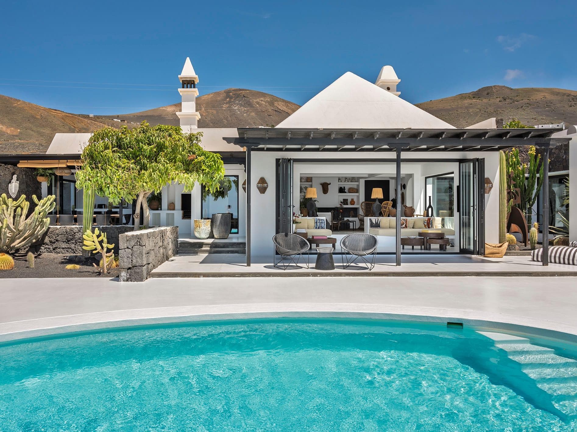 This modern white villa with central courtyard features an inviting pool. It features dark touches, sun loungers, glass doors and desert-inspired landscaping, set against a backdrop of arid hills under partly cloudy skies.