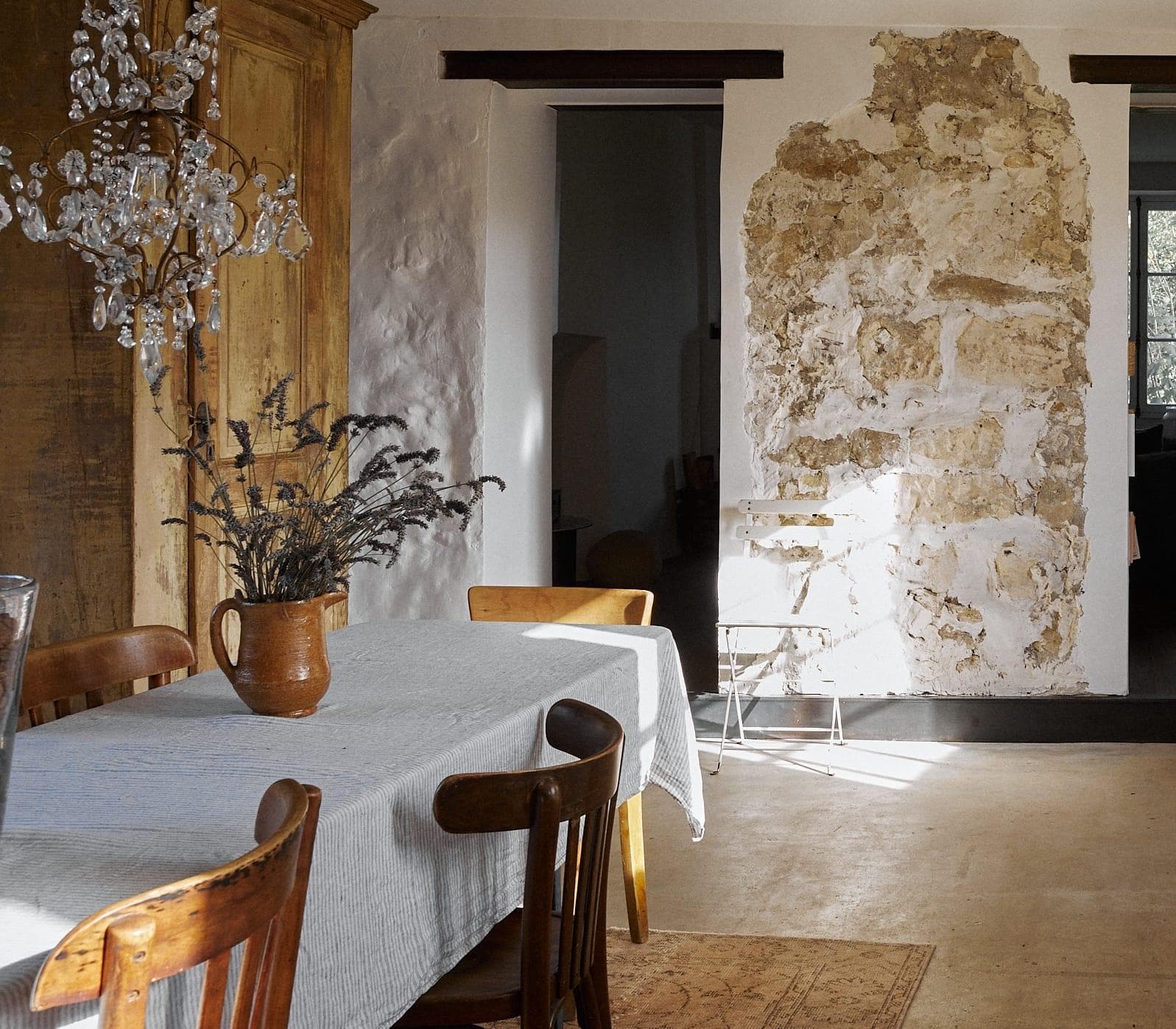 A rustic dining room with a wooden table covered by a gray tablecloth. A ceramic jug with dried plants sits on the table. The room has wooden chairs, a chandelier, and a wall with exposed stonework. Sunlight streams in from a nearby window.