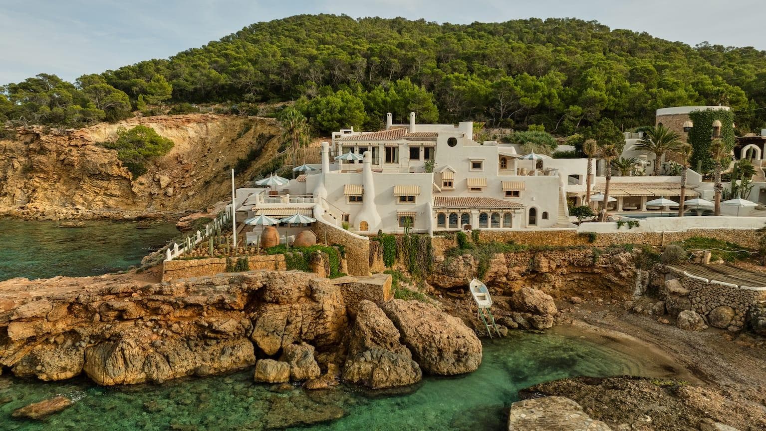 A luxurious white villa stands on a rocky coastal plot, surrounded by lush vegetation. The building overlooks a turquoise sea with stone steps leading down to the water, and tall trees cover the hillside in the background.