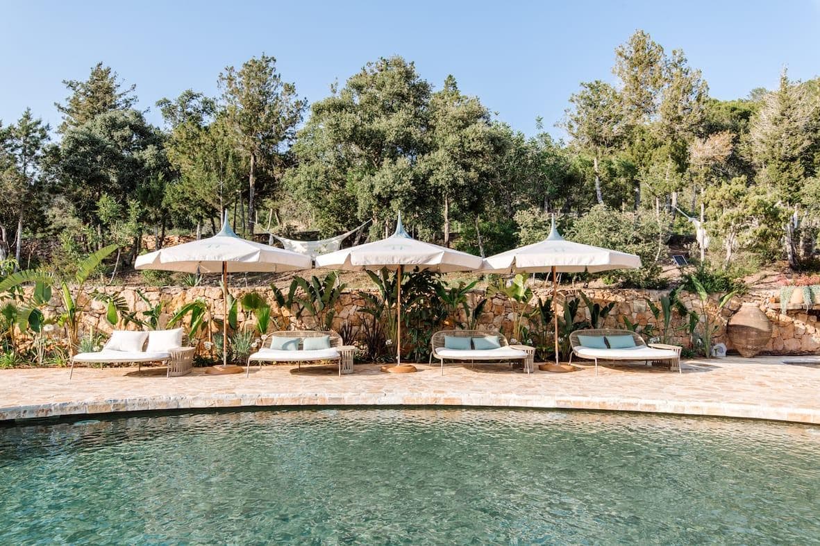 Outdoor pool area with several white parasols and sun loungers on a stone terrace. The pool is surrounded by lush vegetation and trees. The clear, sunny sky reinforces the peaceful atmosphere.