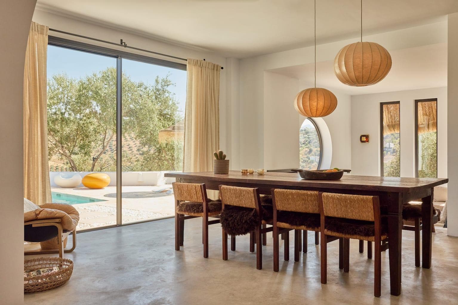 A modern dining room with a wooden table and wicker chairs. Large spherical pendant lights hang above the table. Floor-to-ceiling windows reveal a pool and trees outside, creating an airy and inviting atmosphere.