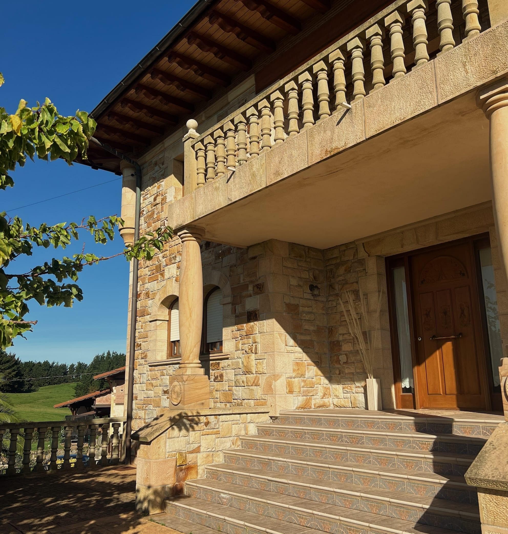 Une maison en pierre avec un escalier carrelé, une porte en bois, des colonnes et des balustrades décoratives. Un arbre et un paysage verdoyant sont visibles sous un ciel bleu clair.