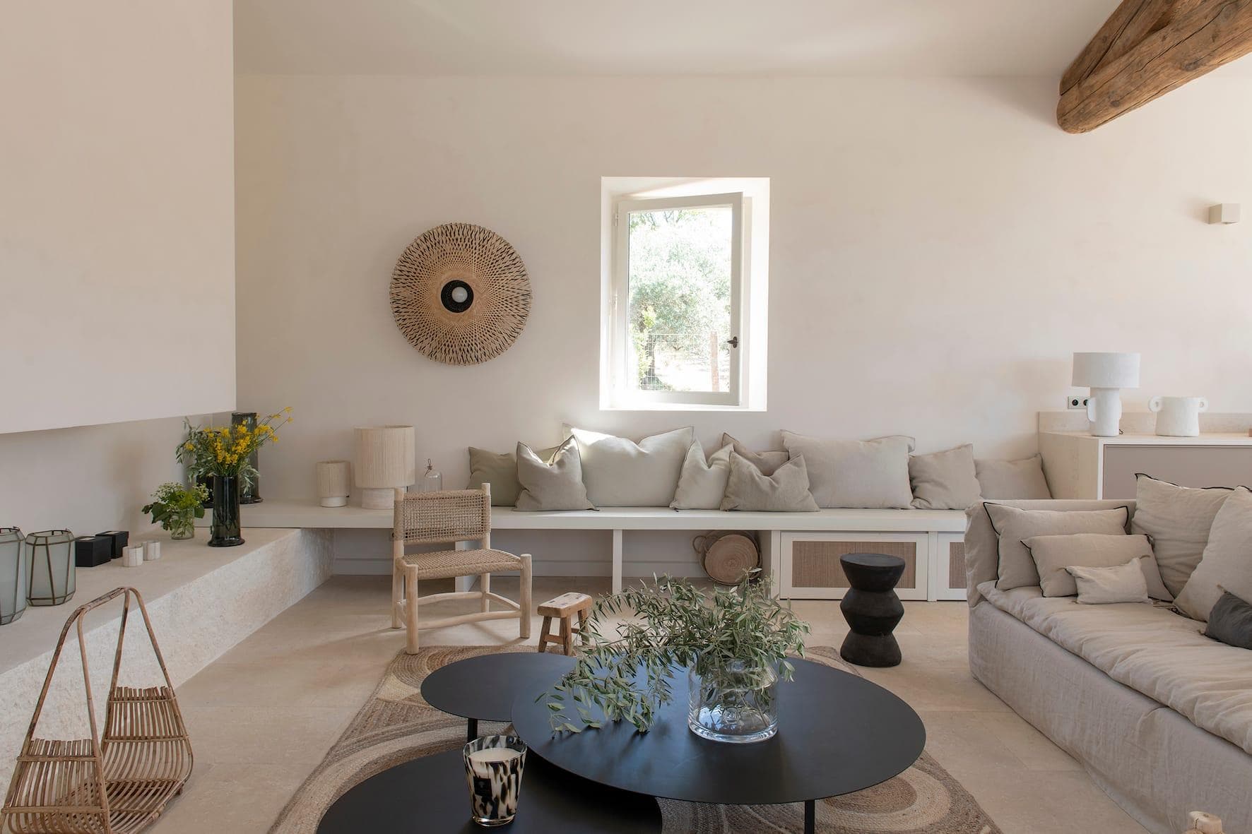 Un salon lumineux et minimaliste avec des murs crème, des touches de bois naturel, une banquette intégrée avec des coussins neutres, un canapé clair, des tables basses rondes noires et des objets décoratifs tissés. La lumière du soleil entre par une petite fenêtre.