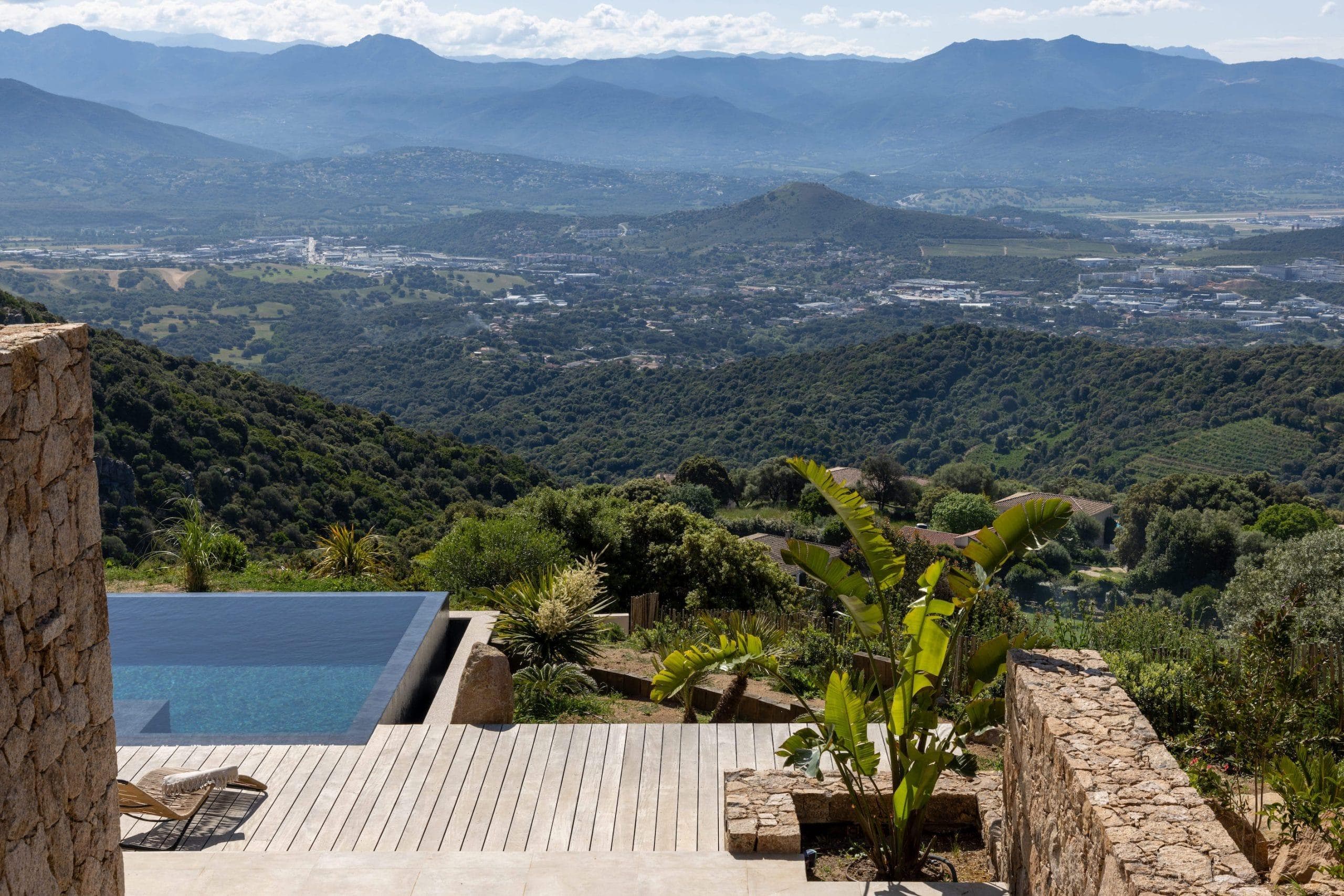 A modern terrace with a small swimming pool overlooks green hills and a distant valley with a town. Stone walls, a wooden deck and tropical plants frame the picturesque mountain landscape under a partly cloudy sky.