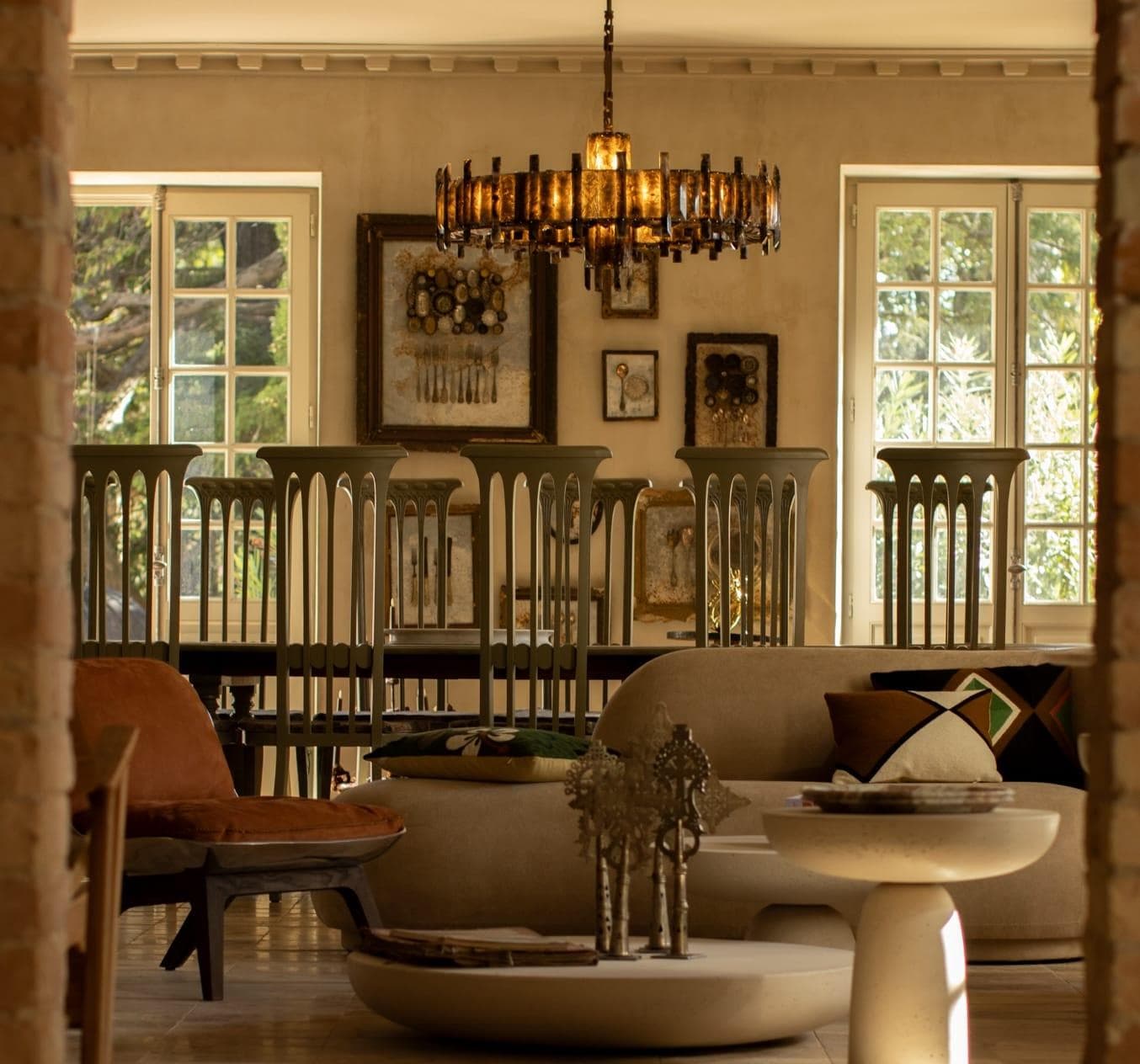 A stylish, sunlit living room features modern furniture, a round coffee table, unique decor, a large chandelier, and framed art on the wall, with tall windows letting in natural light.