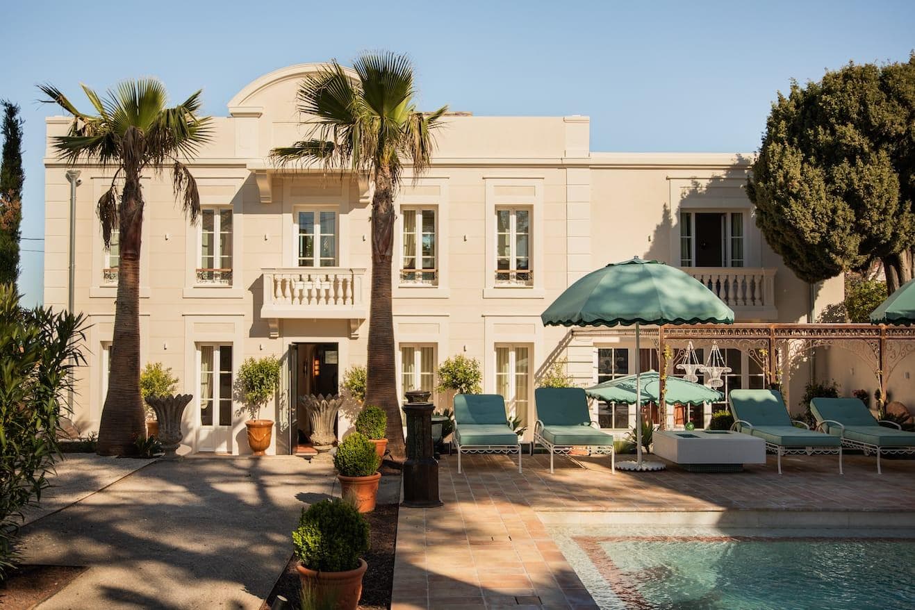 Une villa de deux étages de couleur crème avec des volets verts, des palmiers et des plantes en pot, dispose d'une piscine, de chaises longues bleu sarcelle et de parasols par temps ensoleillé.