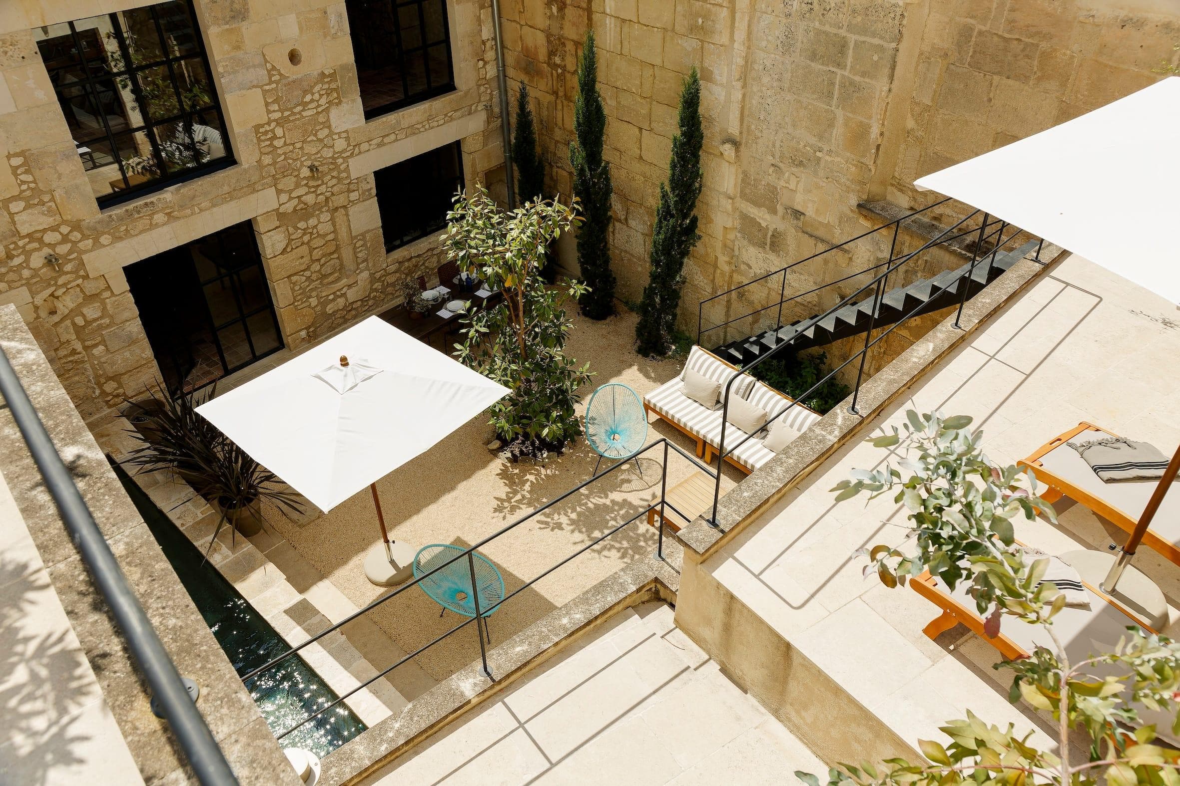 Une cour en pierre ensoleillée dispose d'un parasol blanc au-dessus d'un coin salon avec des coussins rayés, de petites tables rondes, des plantes vertes et des chaises longues à côté d'un bassin réfléchissant étroit.