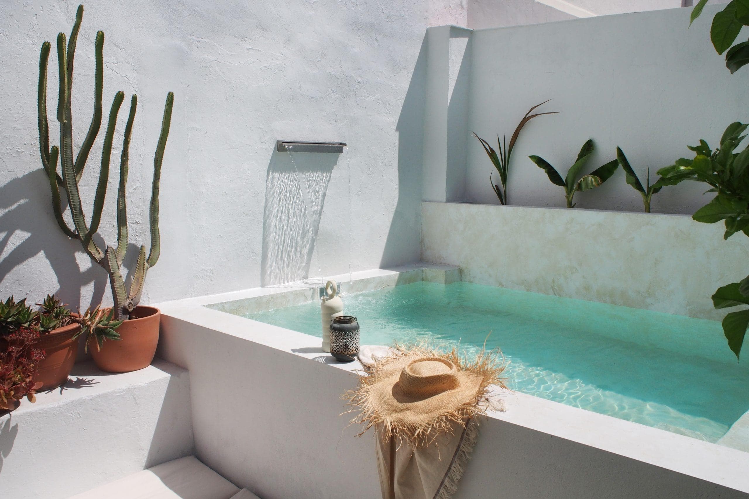 Une petite piscine extérieure avec de l'eau turquoise claire, un chapeau de paille et une serviette sur le bord, des cactus et des plantes en pot à proximité, et une cascade moderne contre des murs blancs.