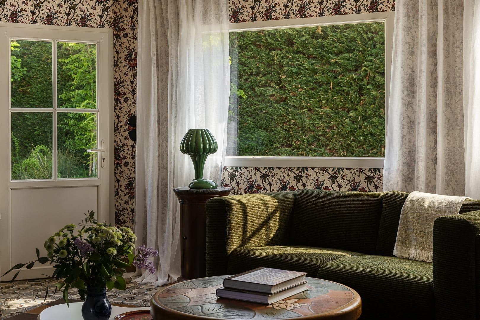 A cozy living room at La Maison du Meunier features a green sofa, floral-patterned wallpaper, and a round table with books and flowers. Soft light filters through sheer curtains, revealing lush greenery outside the window.