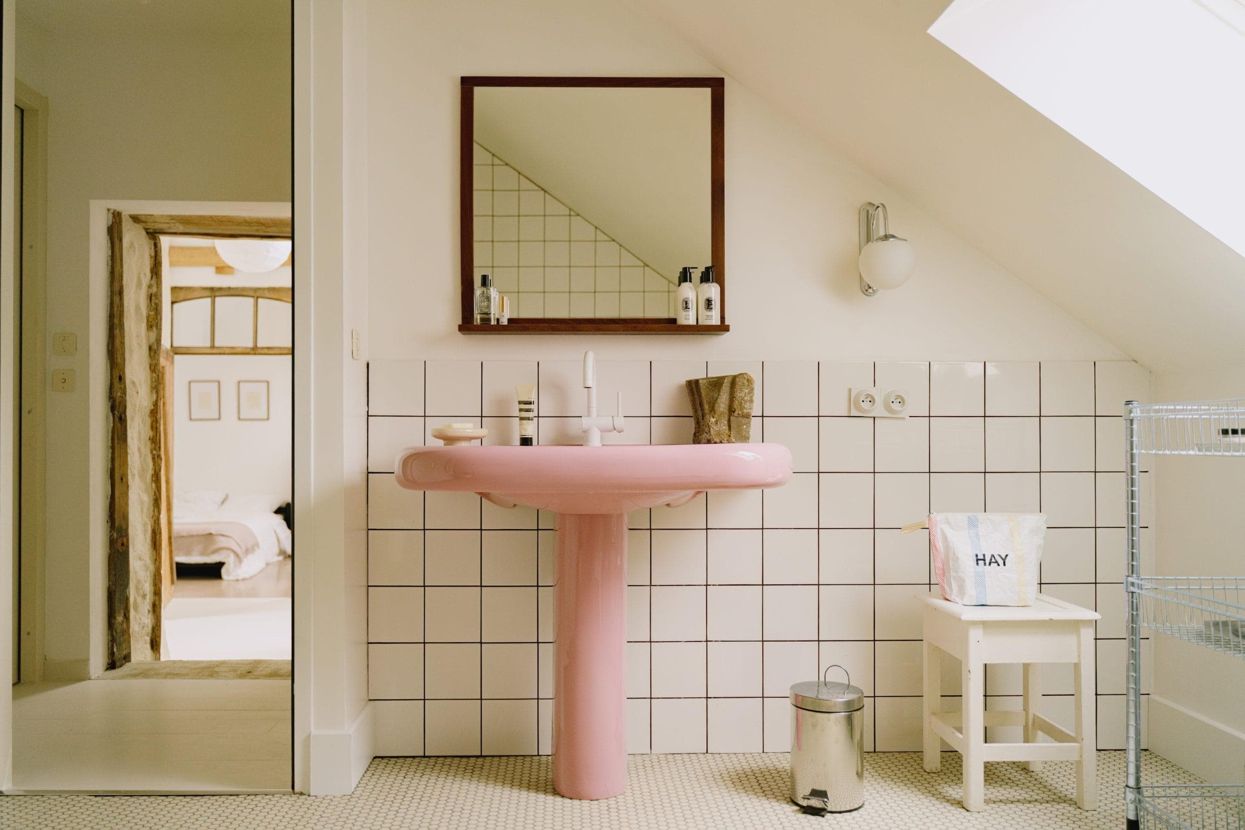 A bathroom with square white tiles and a pink washbasin beneath a wood-framed mirror. A small stool with a bag, a wastebasket and toiletries, bathed in natural light by a sloping skylight.