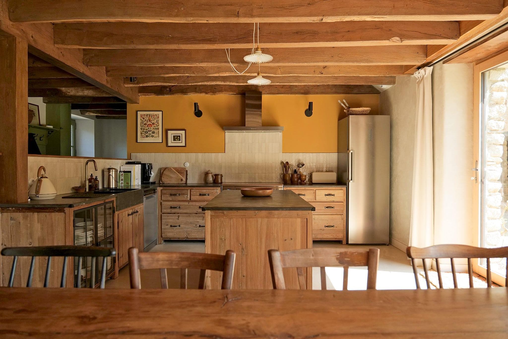 Une cuisine rustique avec poutres apparentes, placards en bois, îlot central et réfrigérateur en inox. La lumière du soleil pénètre par une fenêtre, illuminant cet espace chaleureux et cosy. Quatre chaises en bois sont disposées autour d'une table à manger au premier plan.