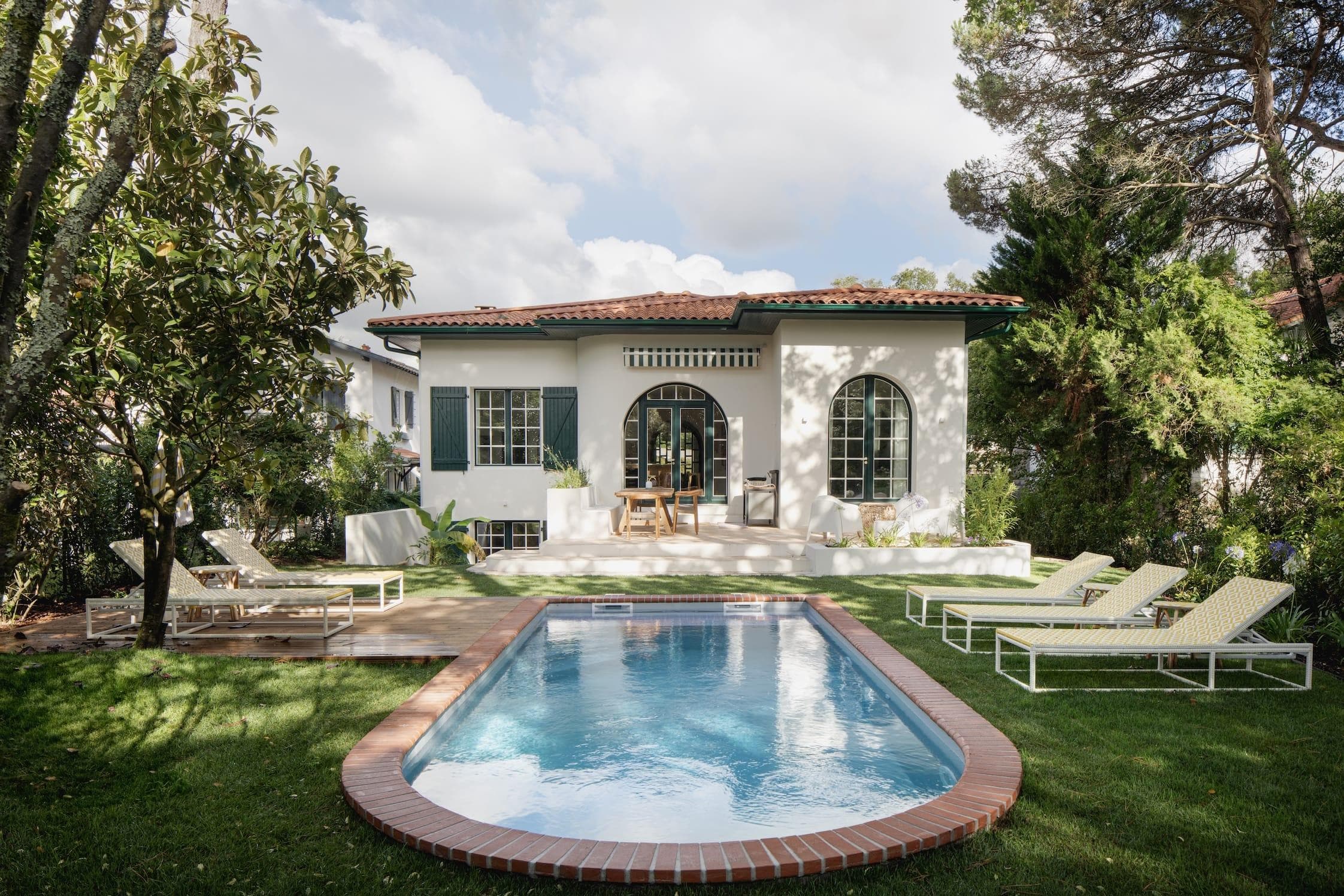 A small rectangular brick-lined swimming pool sits in a lush green garden, surrounded by sun loungers. Behind it is a white house with arched windows, a tiled roof and a shaded patio with a dining table. Trees and greenery decorate the setting.