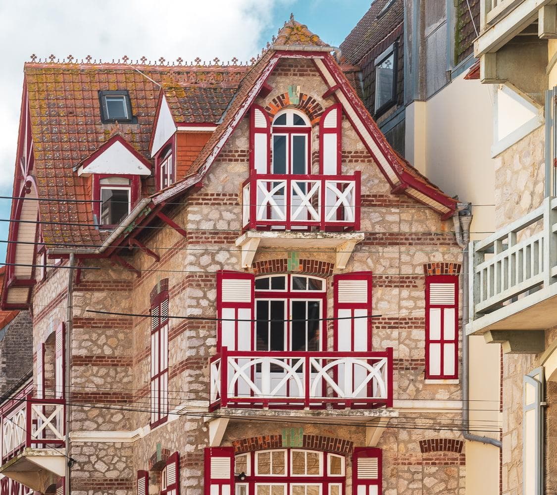 A charming stone building with red and white wooden trim, balconies, and shuttered windows, featuring a steep tiled roof and intricate architectural details.