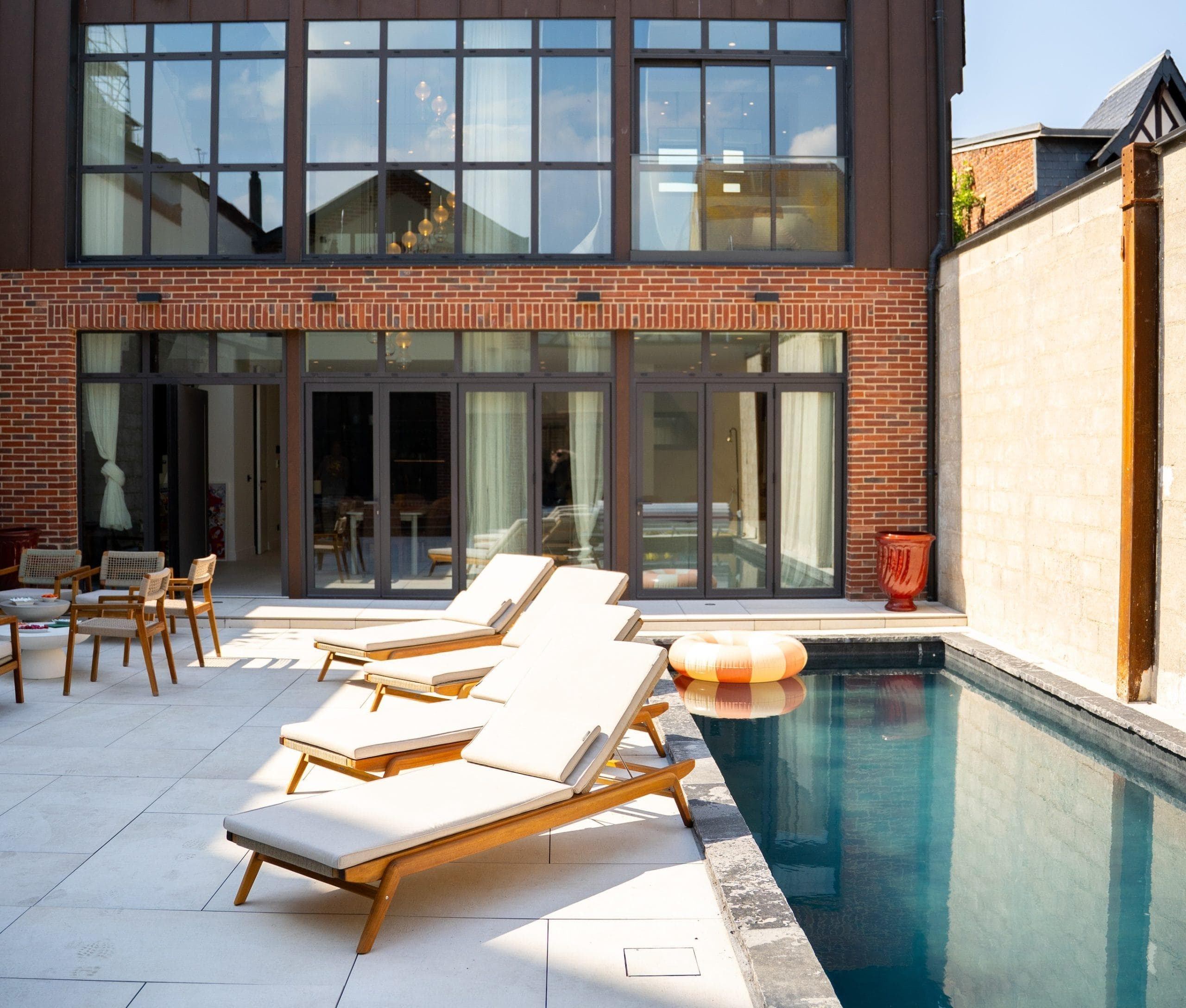 Four sun loungers and a float line the edge of a modern outdoor swimming pool. Behind the pool, large windows and sliding doors open onto a contemporary brick and glass building.