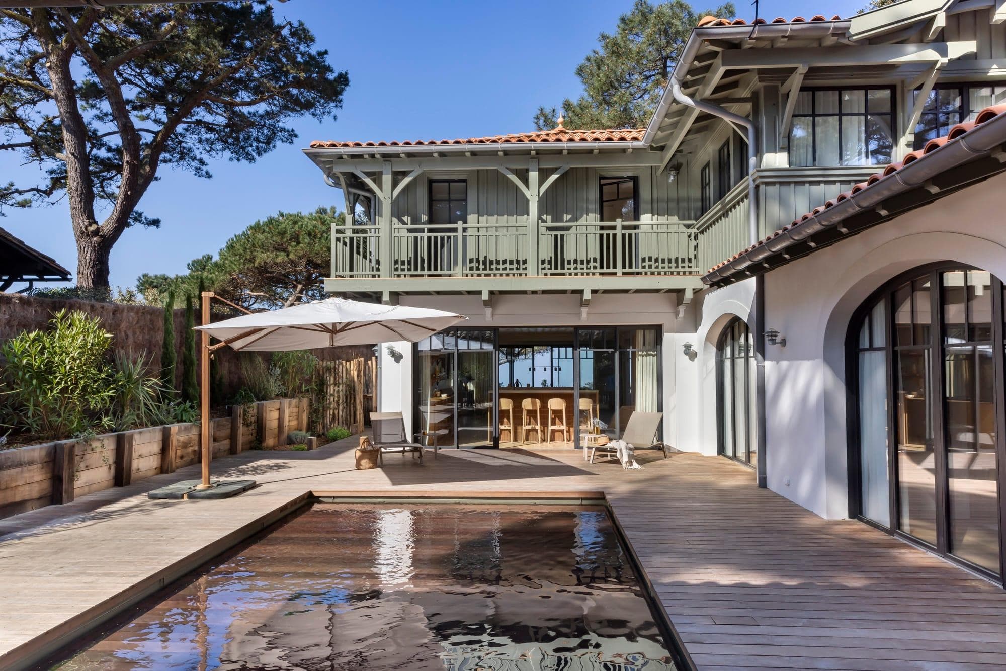 Maison moderne à deux étages avec extérieur vert et blanc, balcons en bois, grandes fenêtres et portes coulissantes s'ouvrant sur une terrasse en bois avec une petite piscine, un parasol, des sièges de bar et des chaises longues, entourée d'arbres.