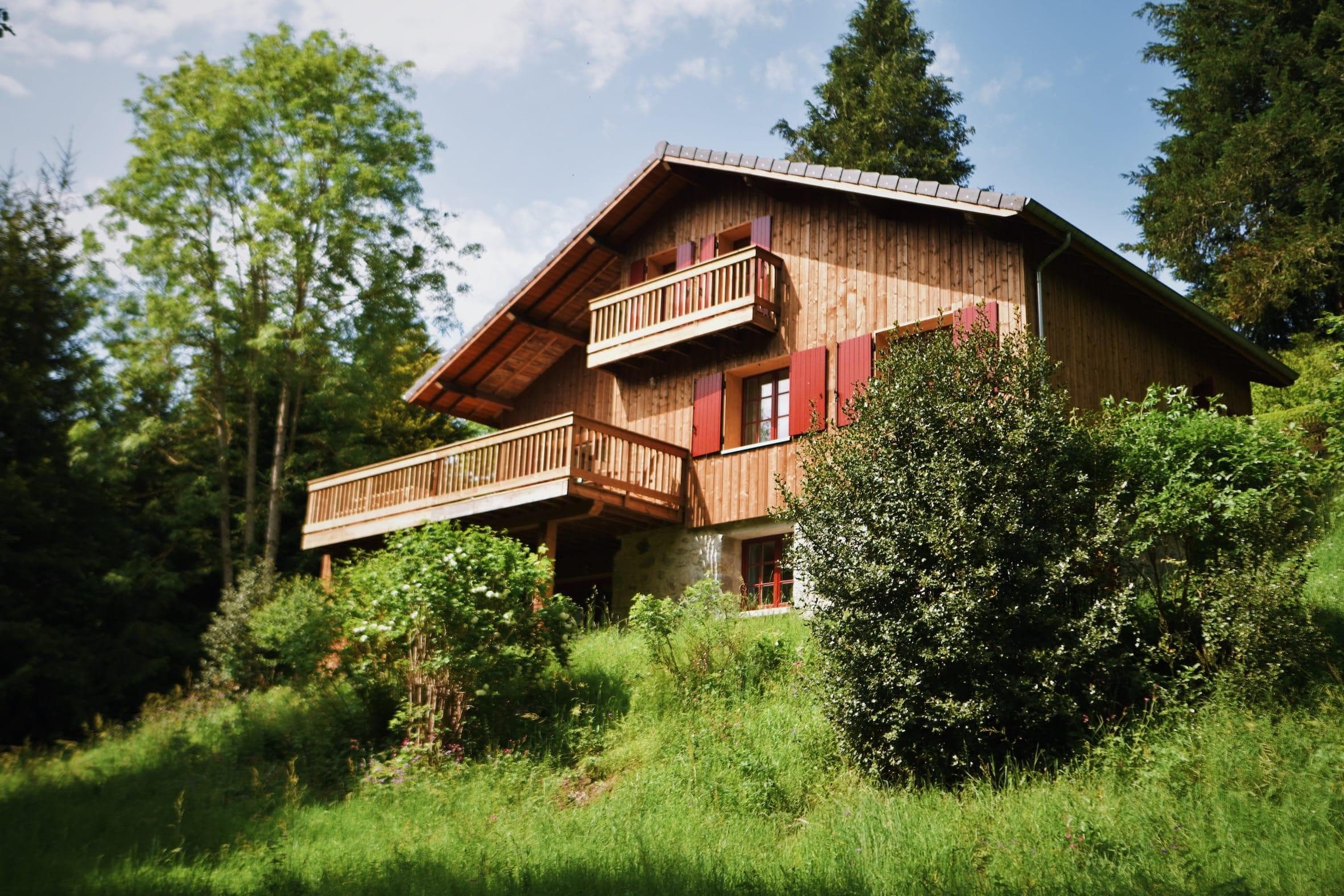 A wooden hut with a balcony and red shutters is nestled among lush green trees and bushes on a grassy hillside under a partly cloudy sky.