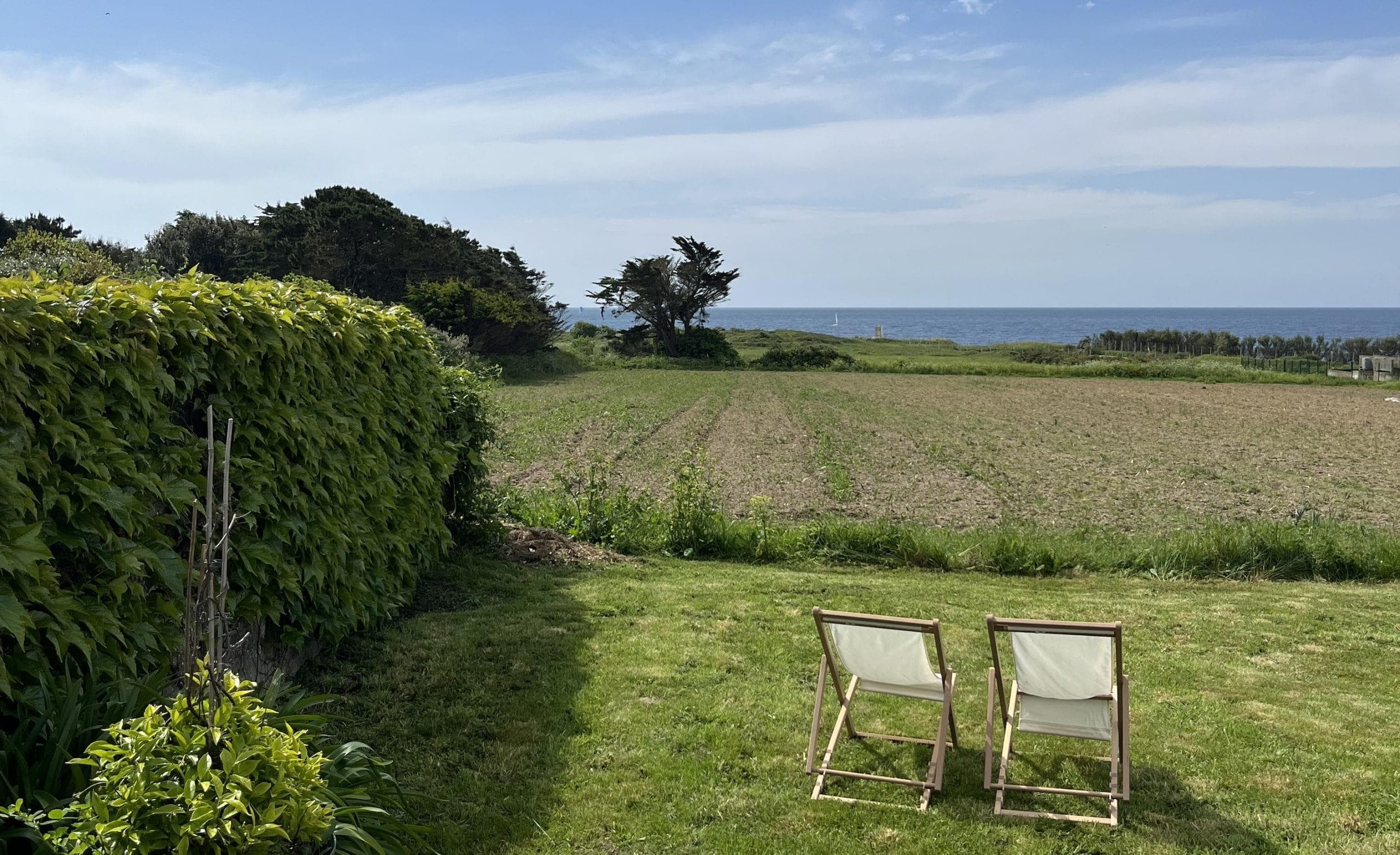 Two empty wooden chairs sit on a lawn, facing a ploughed field and the ocean. Hedges and green trees line the stage under a partly cloudy blue sky.