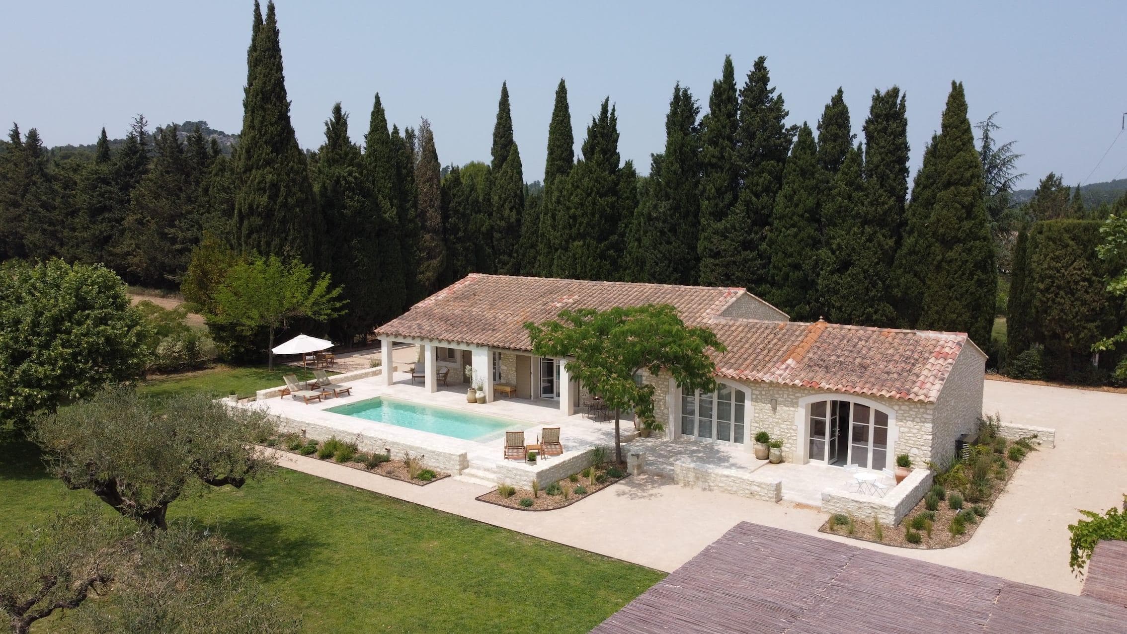 Vue aérienne d'une villa rustique en pierre avec un toit en tuiles rouges, une piscine, des chaises longues et des parasols, entourée de pelouses vertes, de grands arbres et de jardins paysagers.
