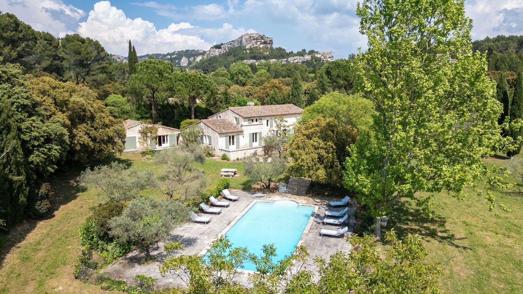 Une grande villa avec un patio en pierre et une piscine entourée de chaises longues, située au milieu d'arbres et de verdure avec une colline rocheuse et un village en arrière-plan sous un ciel partiellement nuageux.