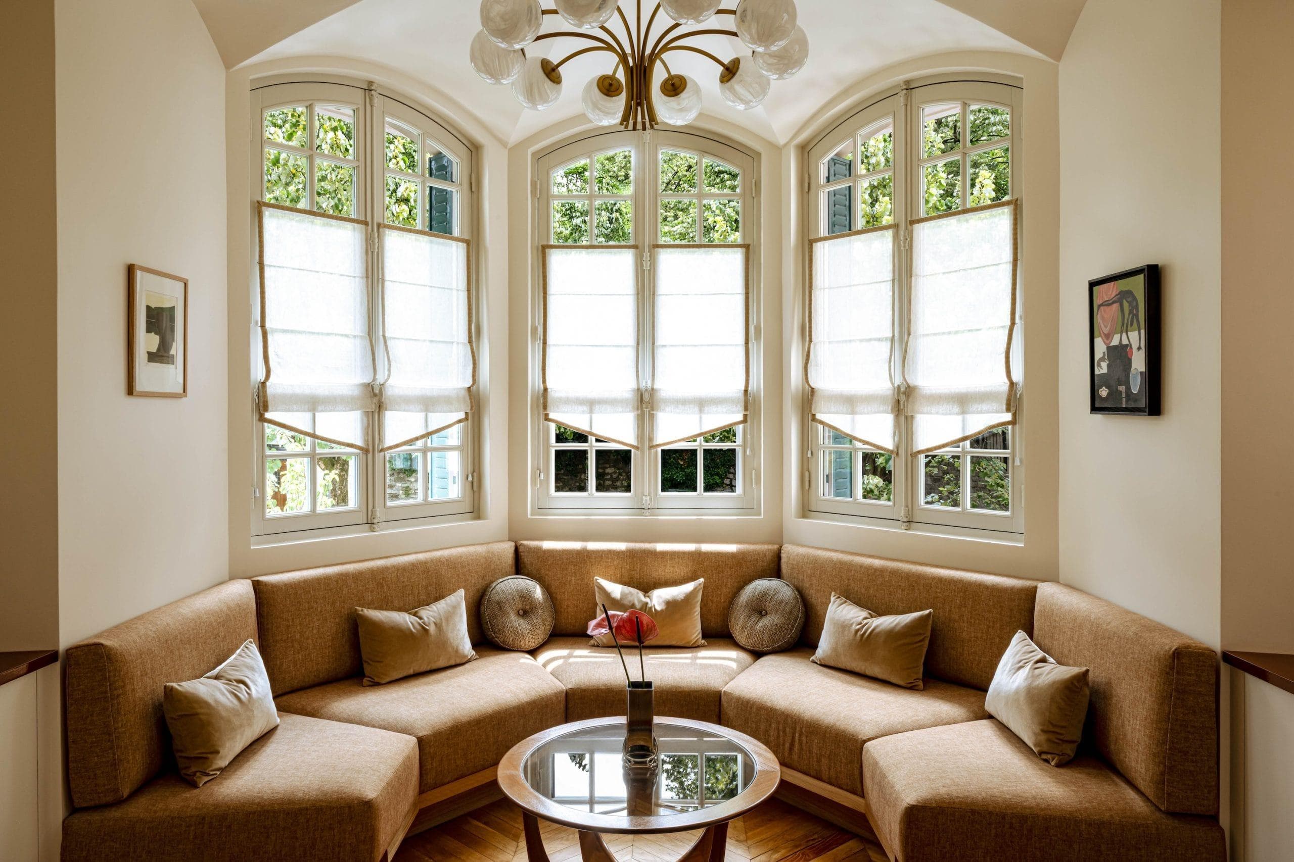 A cozy corner seating area inspired by Claves architecture, with beige cushions, round pillows, and a glass coffee table with a single flower vase. Large windows with white blinds fill this Villa Junot space with natural light as a chandelier glows overhead.