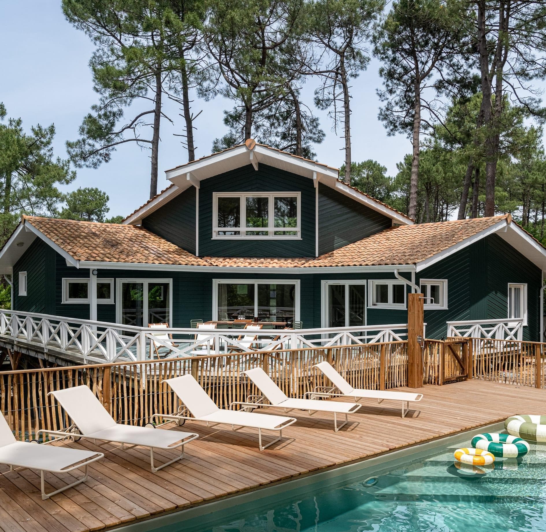 Une maison moderne de style cabane, avec un bardage vert foncé et des bordures blanches, se trouve au milieu de grands pins, avec une spacieuse terrasse en bois, des chaises longues blanches et une piscine au premier plan. Des flotteurs de piscine gonflables sont dans l'eau.