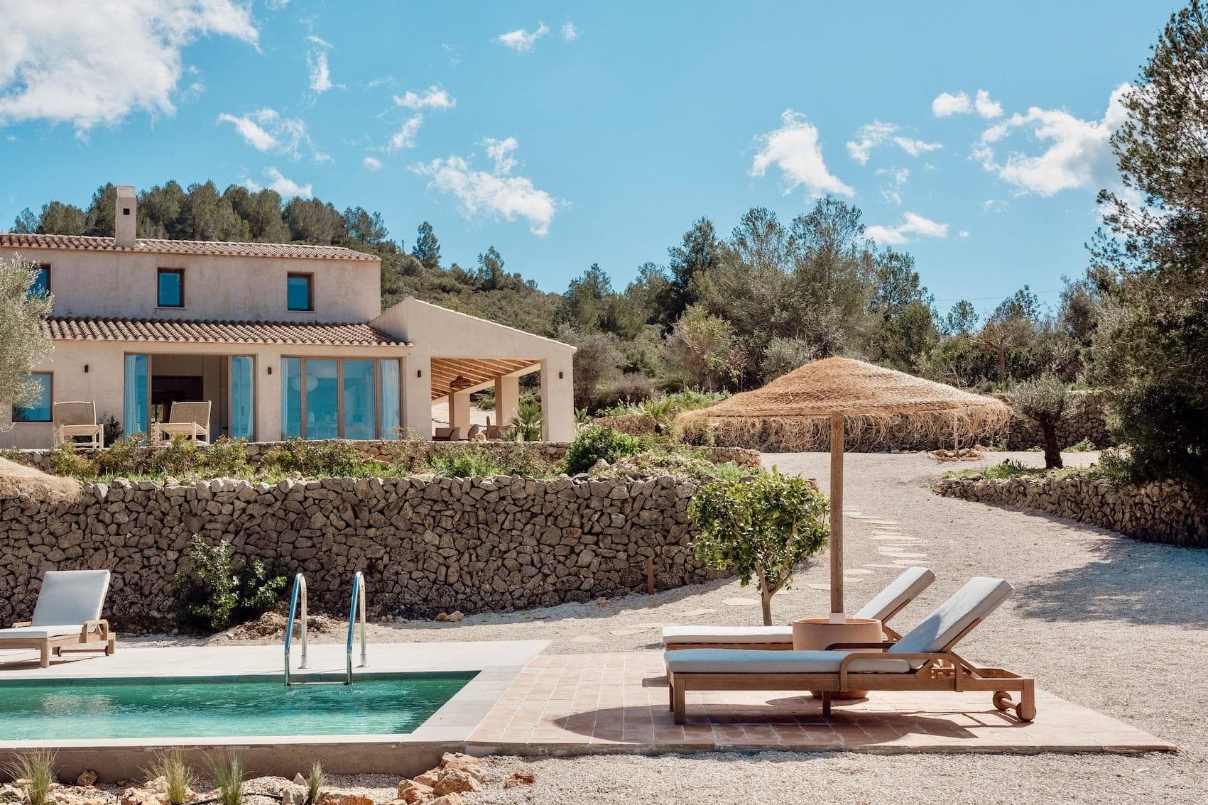 A stone-walled villa with large windows stands among the trees, with a small swimming pool and two sun loungers in the foreground. A thatched parasol offers shade nearby, under a blue sky dotted with clouds.
