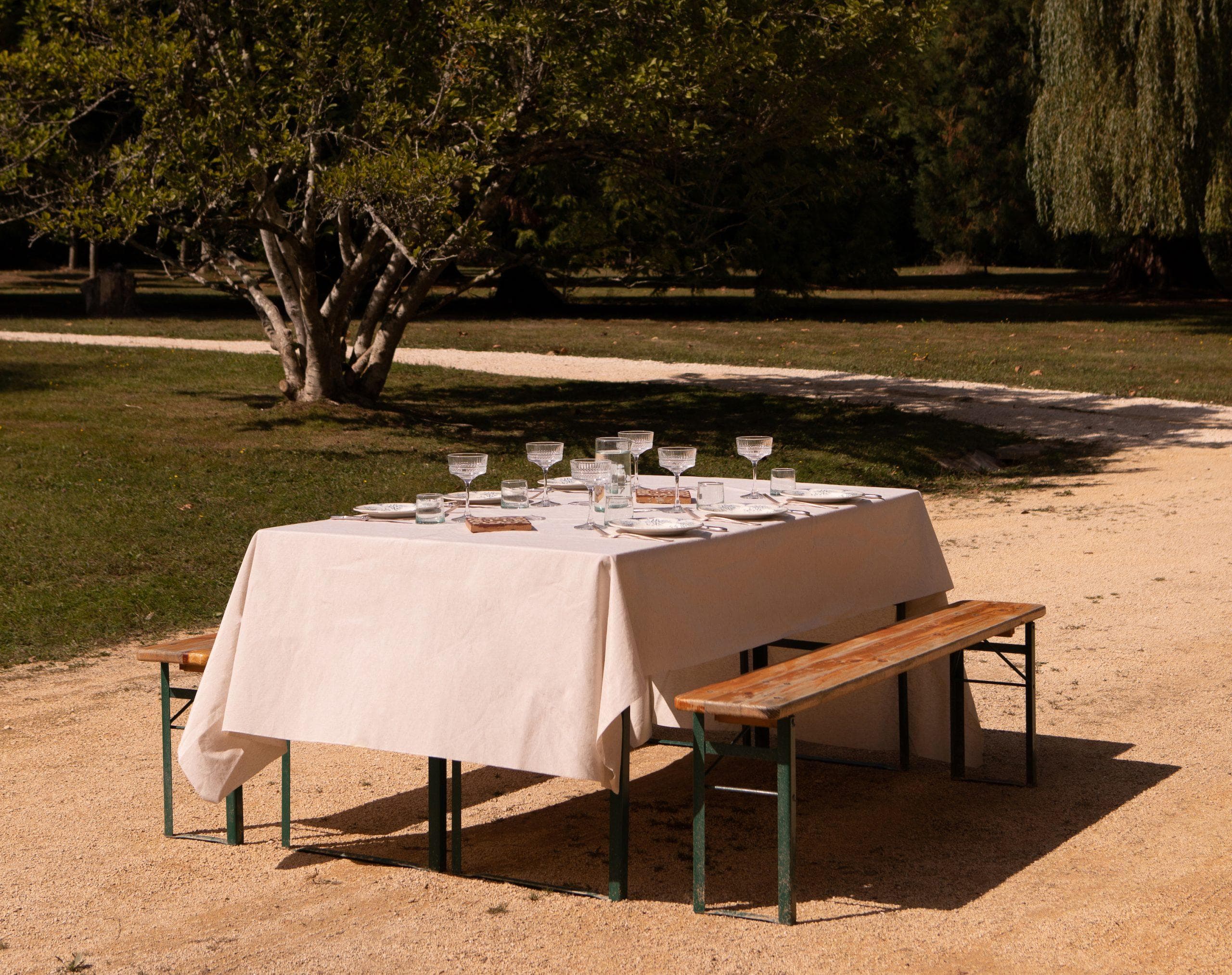 Une table de pique-nique en bois avec une nappe blanche est installée à l'extérieur, avec des verres et des couverts pour six personnes. Elle orne un chemin de gravier entouré d'herbe, d'arbres et d'ombres sous un ciel ensoleillé - parfait pour ceux qui recherchent le charme des « maisons de campagne à louer ».