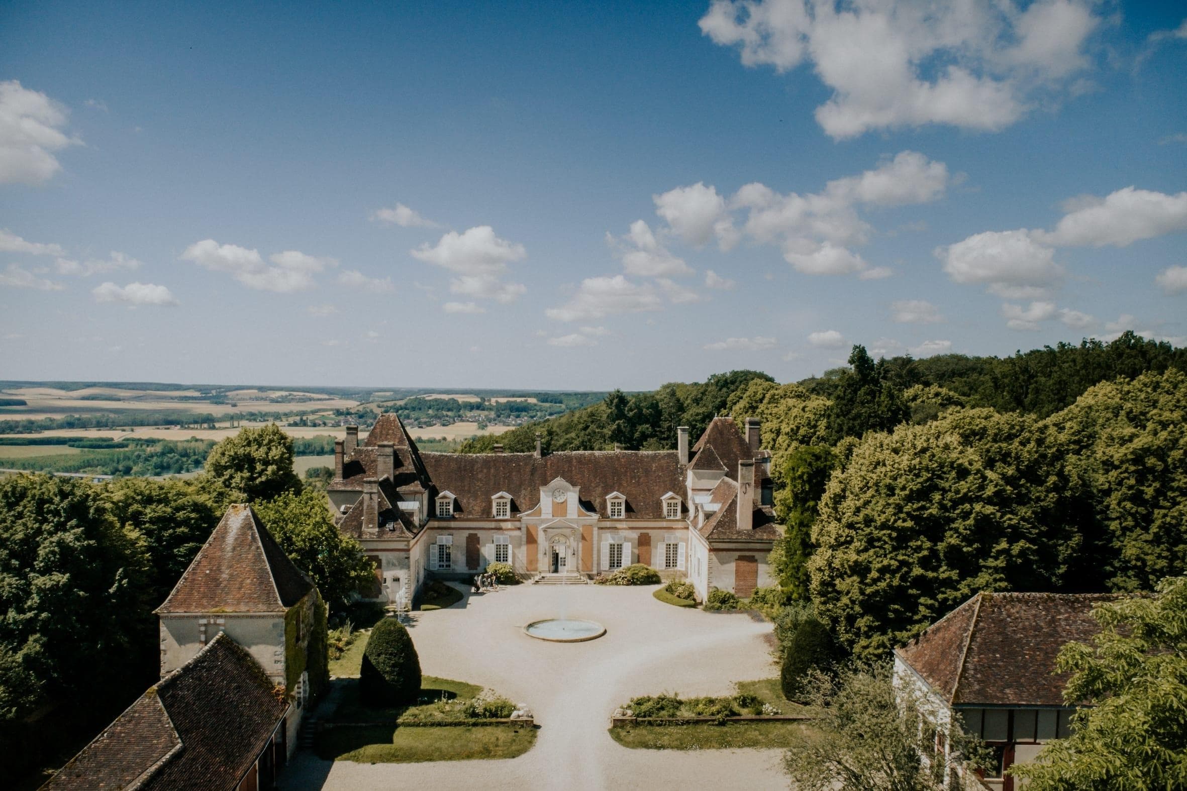 Vue aérienne d'un château historique entouré d'arbres luxuriants et de campagne. Le bâtiment présente des façades en pierre, des toits triangulaires et une allée circulaire avec une fontaine au centre. Le ciel au-dessus est bleu avec des nuages épars.