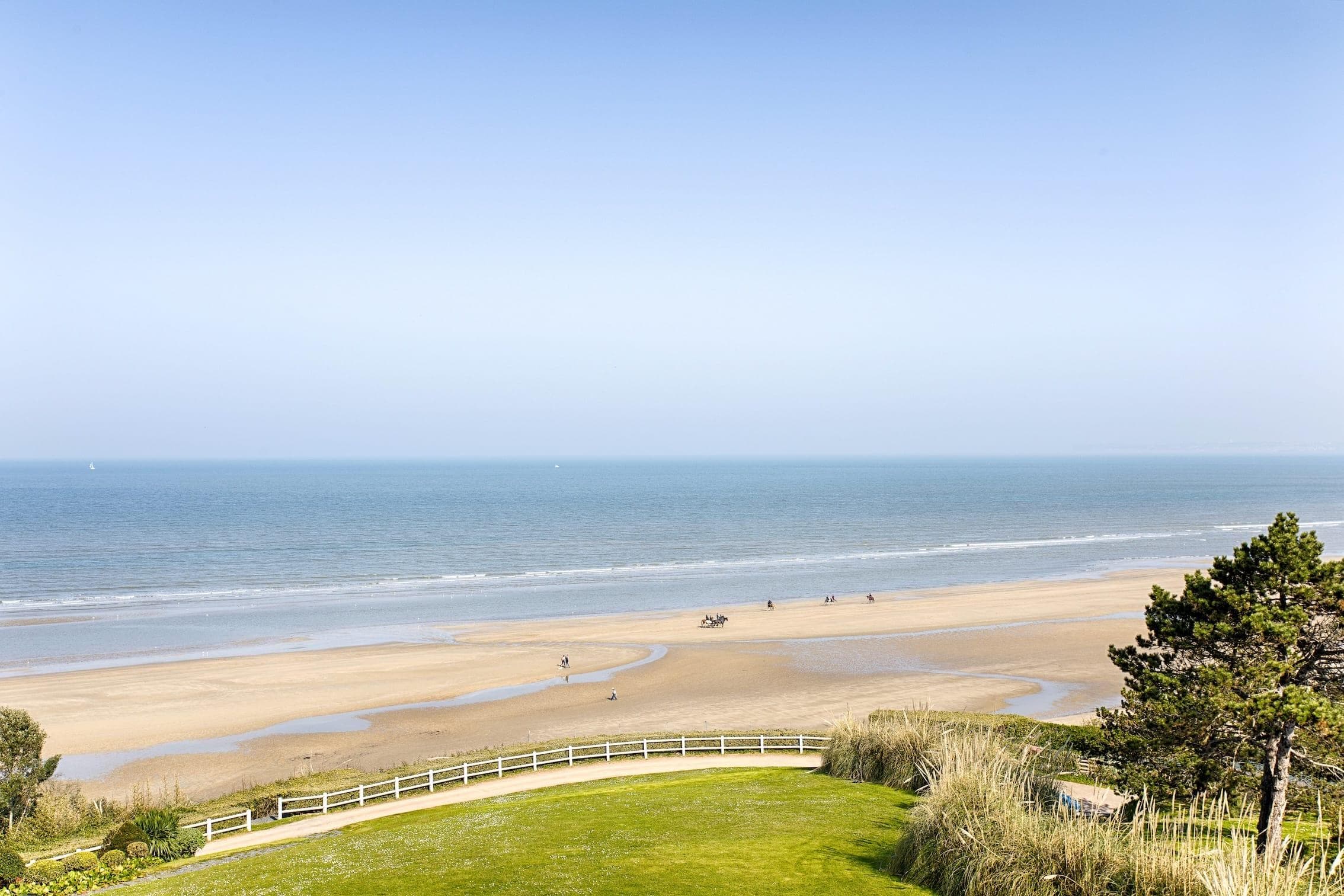 Vue d'une plage de sable avec plusieurs personnes et chevaux au loin. L'océan est calme et une zone herbeuse bordée d'une clôture blanche apparaît au premier plan. Le ciel est clair et bleu.