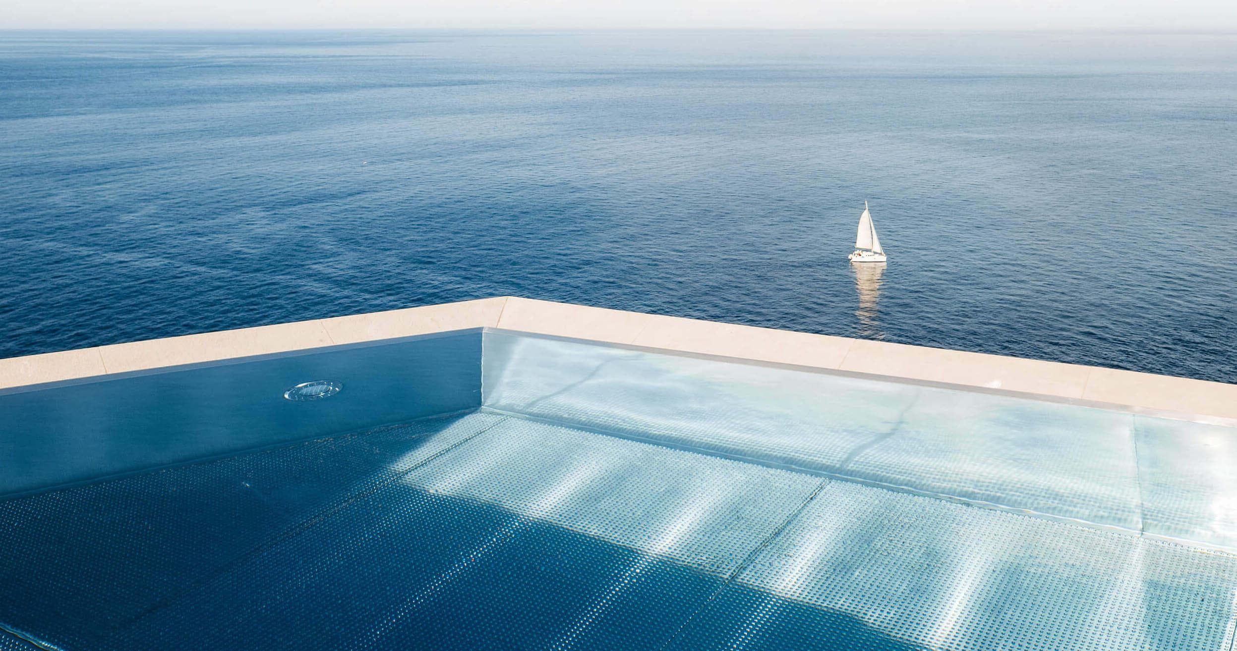 Une piscine à débordement aux eaux bleues et limpides surplombe un océan vaste et calme. Au loin, un voilier blanc glisse sur l'eau. L'horizon se brouille là où le ciel rencontre la mer, créant une scène sereine et paisible.