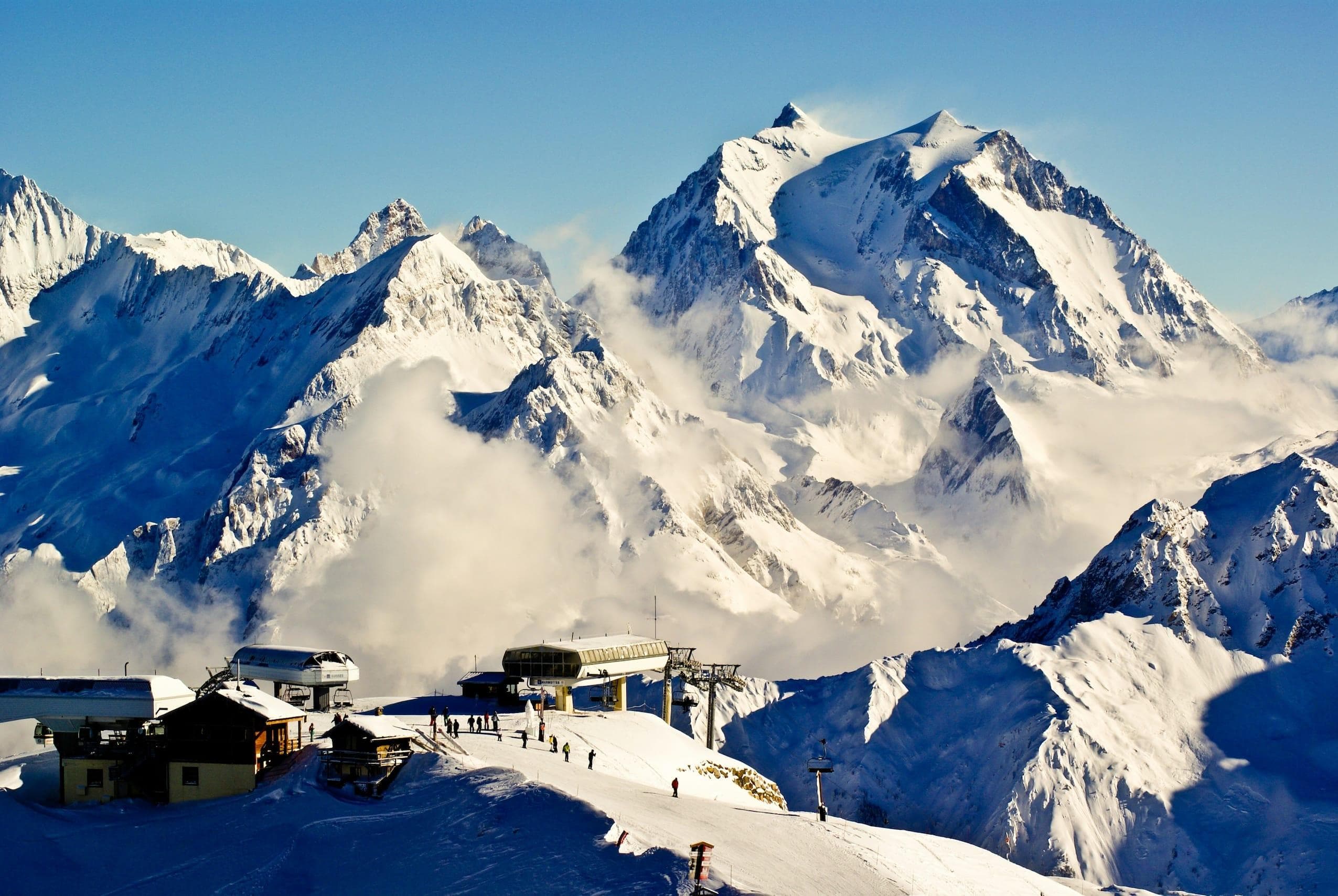 Des montagnes enneigées sous un ciel bleu clair, avec des skieurs sur une piste près des stations de télésiège au premier plan. Le brouillard flotte dans les vallées sous les sommets imposants.