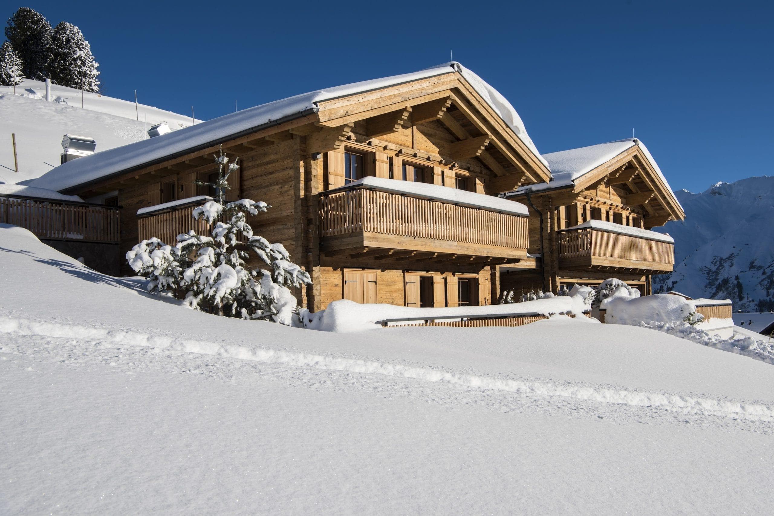 A picturesque snowy scene features two wooden cabins with balconies nestled on a snow-covered hillside under a clear blue sky. Snow-laden trees add to the serene, wintry atmosphere.