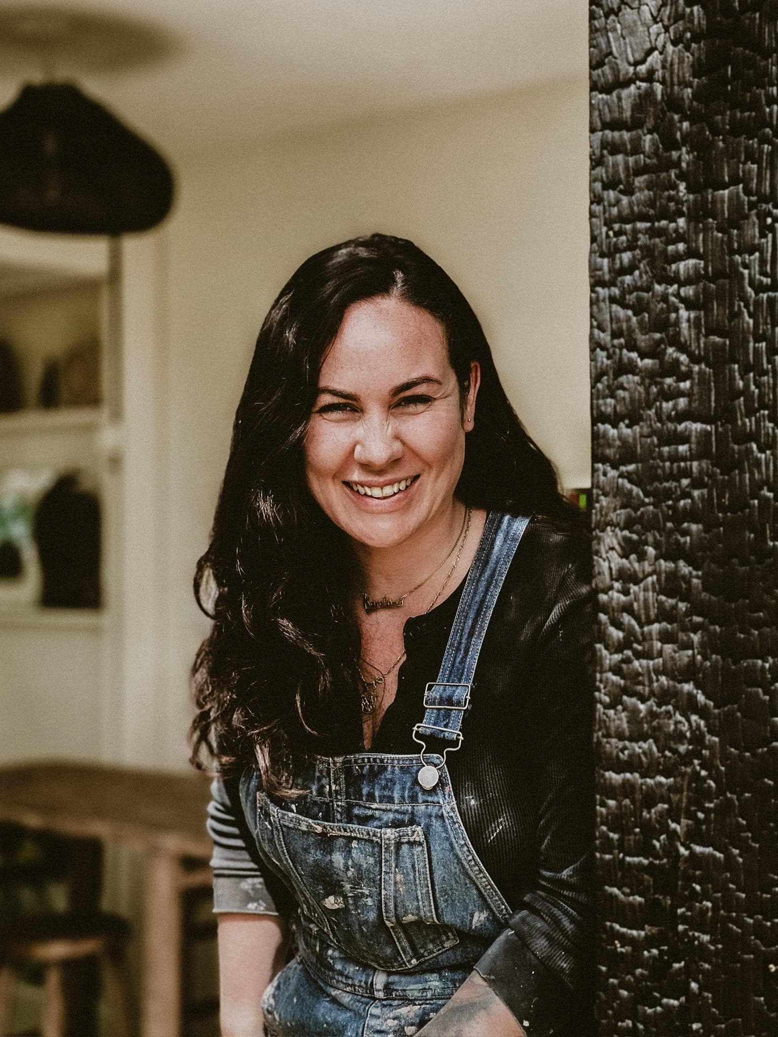 Une femme aux longs cheveux noirs sourit en se tenant dehors. Elle porte une salopette en jean et un haut foncé. L'arrière-plan comprend une table en bois et une porte.