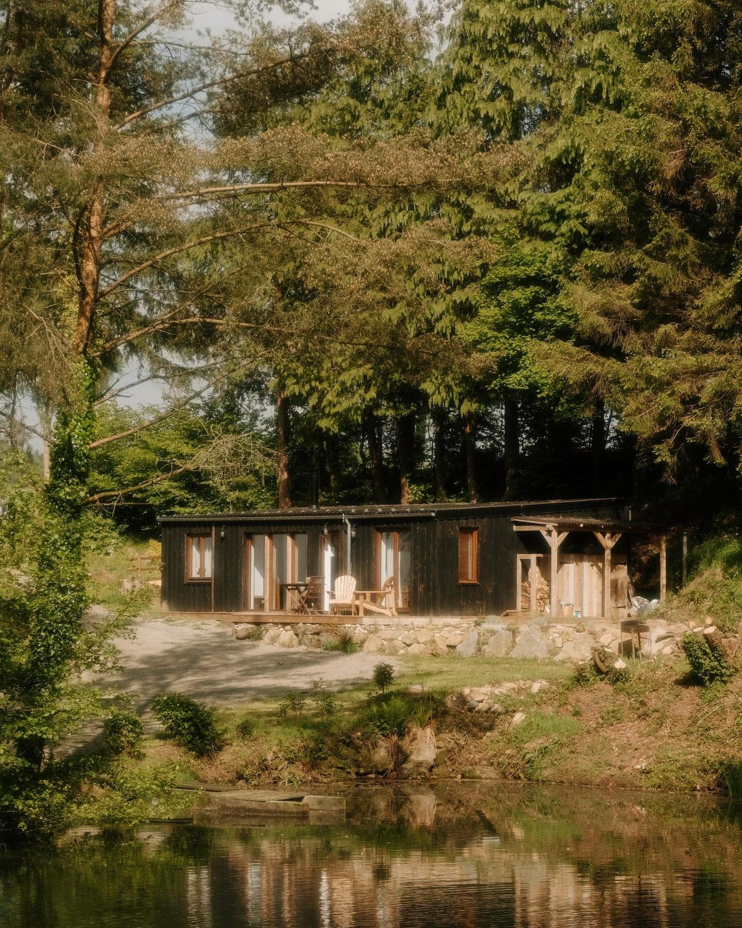 A small, rustic cabin with a wooden exterior is nestled among tall trees by a calm pond. There's a chair and small table on the porch, and sunlight filters through the lush greenery, creating a peaceful, serene atmosphere.
