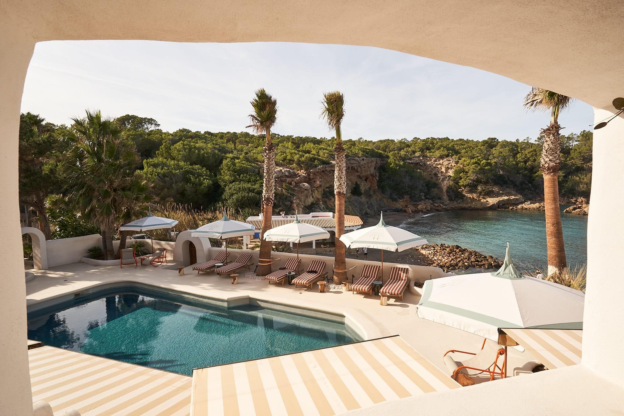 View from a terrace overlooking a swimming pool surrounded by sun loungers and parasols. Beyond the pool, palm trees offer panoramic views of a rocky coastline and calm sea under clear skies.