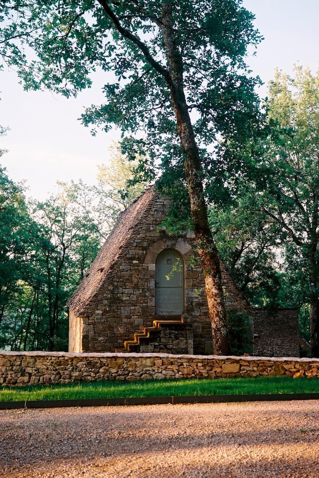 Une petite maison en pierre au toit triangulaire, conçue par Sapiens Architectes, est nichée au milieu de grands arbres. On y accède par un petit escalier en pierre. La lumière du soleil filtre à travers la verdure, créant des ombres tachetées sur le sol. Un mur de pierre court devant.