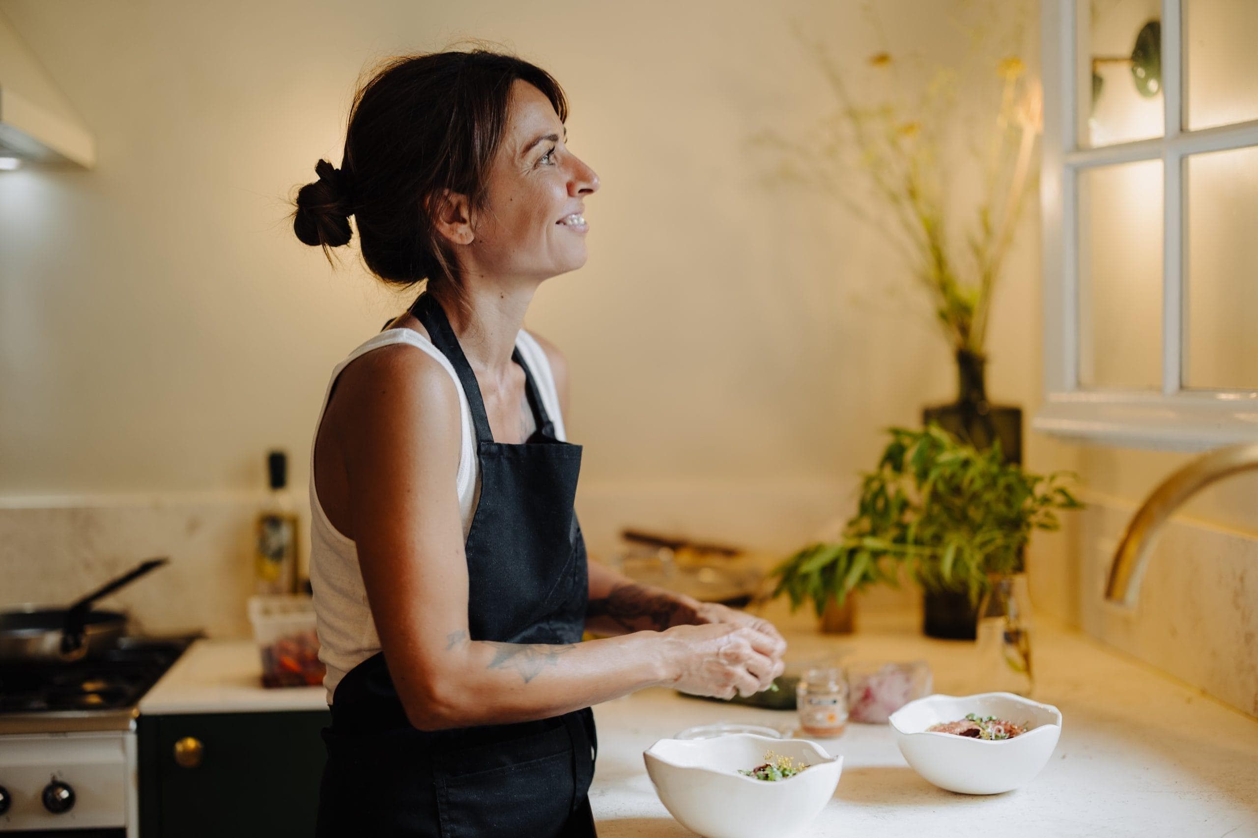 Une femme vêtue d'un tablier noir Maison Mastrorelli et d'un débardeur blanc prépare un repas dans une cuisine, souriante et regardant de côté. Deux bols remplis d'ingrédients sont posés sur le comptoir, avec des herbes et des ustensiles de cuisine en arrière-plan.