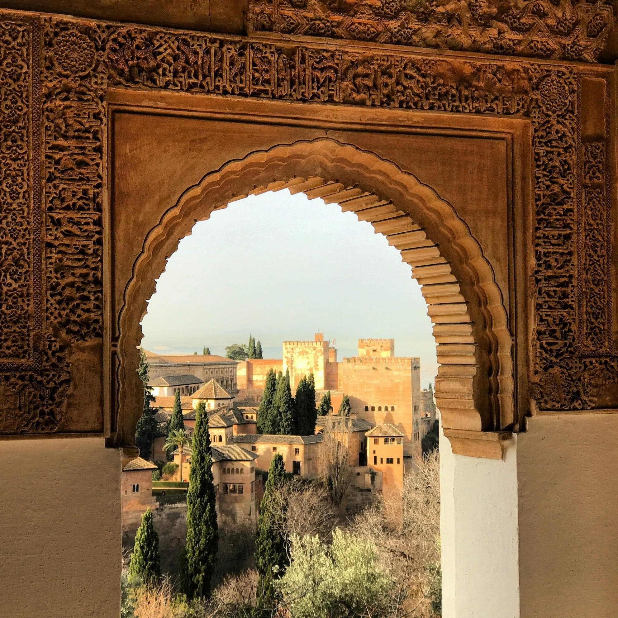 Vue à travers une fenêtre cintrée ornée, mettant en valeur le complexe de l'Alhambra, entouré d'arbres luxuriants, sur fond de ciel dégagé. Les détails complexes de l'arche encadrent magnifiquement l'architecture historique.