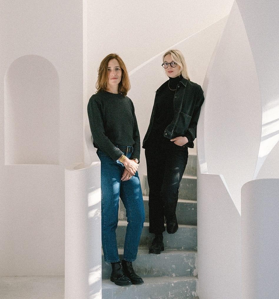 Deux femmes se tiennent sur un escalier incurvé dans un espace lumineux et minimaliste aux murs blancs et aux détails architecturaux en arc. L'une porte un jean et un pull foncé ; l'autre est entièrement vêtue de noir et porte des lunettes.
