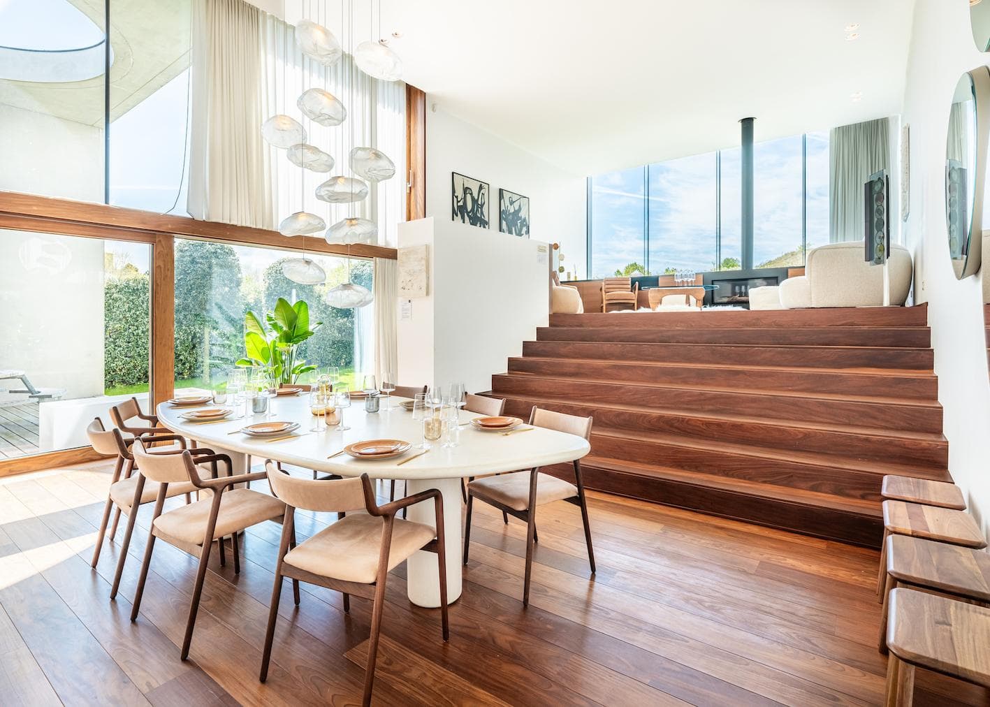 A modern, bright dining room with a white oval table, wooden chairs, and large windows. Wooden steps lead up to a sunlit living area with light-colored furniture and artwork on white walls.