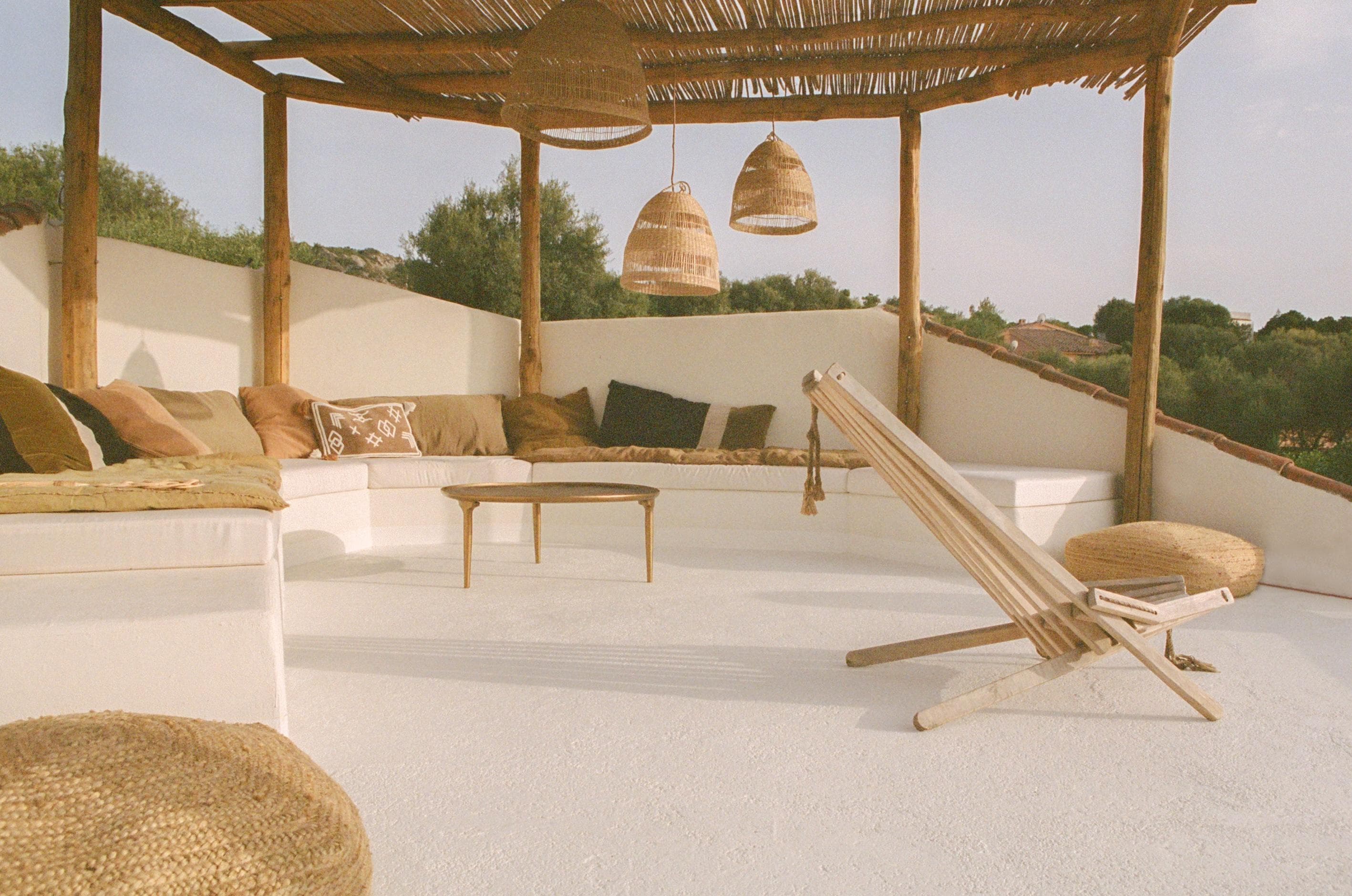 A cozy outdoor lounge with white seating, brown and beige cushions, a wooden coffee table, woven pendant lights, and a folding chair under a wooden pergola, surrounded by greenery and soft natural light.