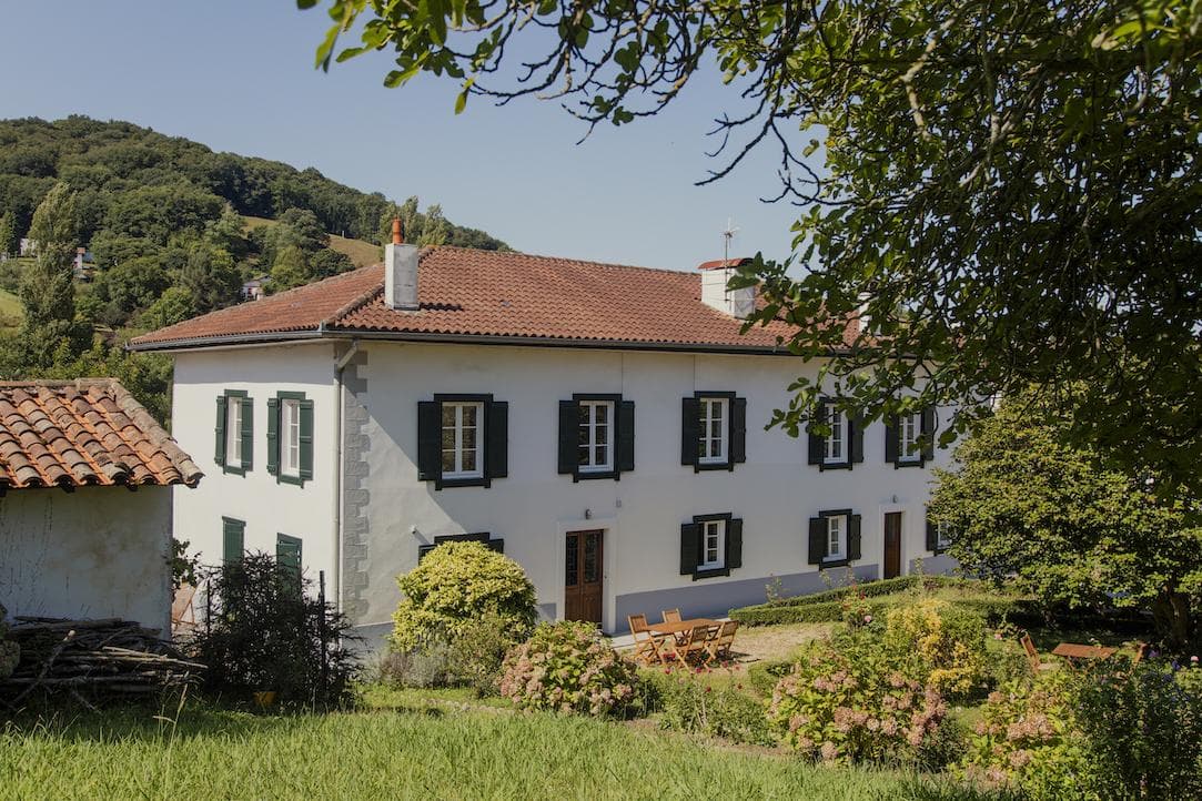 A large white house with dark green shutters sits in a lush garden with wooden outdoor furniture, surrounded by trees and hills under a clear blue sky.