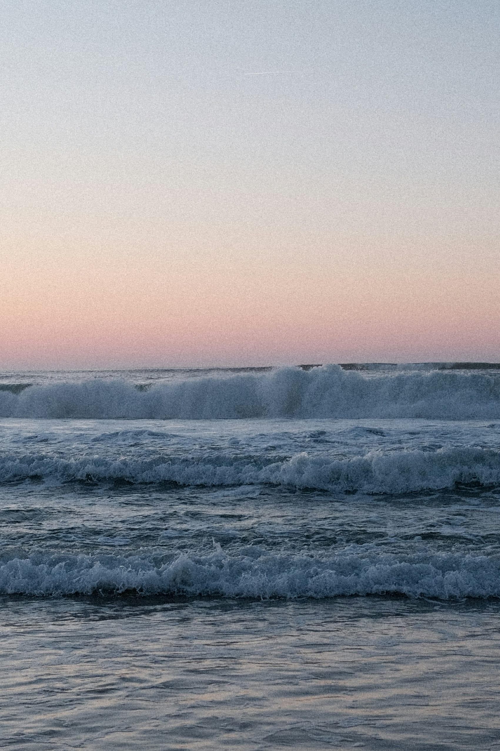 Des vagues déferlant sur le rivage sous un ciel aux couleurs pastel au coucher du soleil, avec de douces teintes roses et bleues se reflétant sur l'eau.