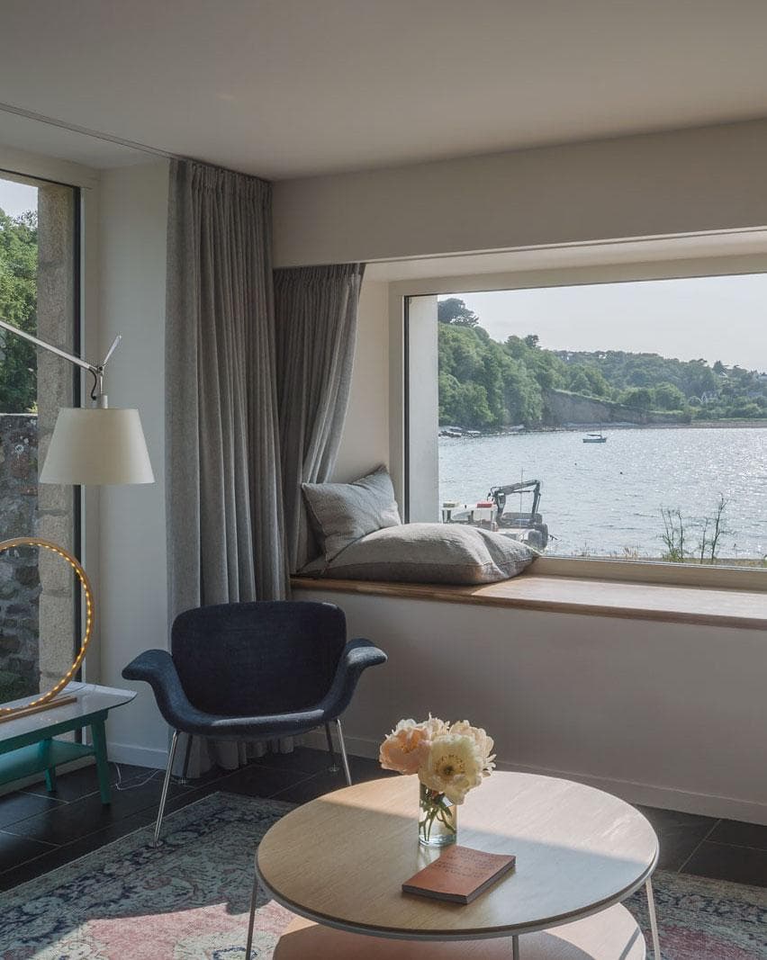 Modern living room with a large window seat overlooking a calm river. Cozy chair, round coffee table with flowers and a book, and grey curtains frame the scenic outdoor view of trees and water.