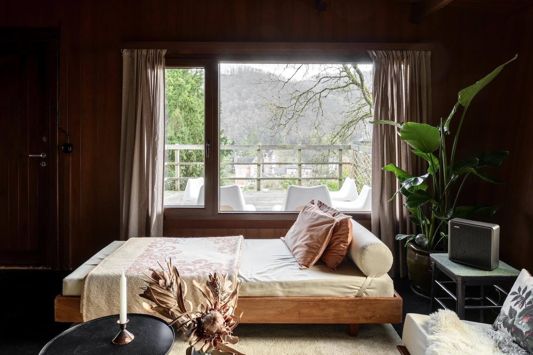 A cozy living room with wood-paneled walls features a daybed with pillows and a blanket, large window with a view of trees and mountains, potted plant, and a small table with dried flowers and a candle.