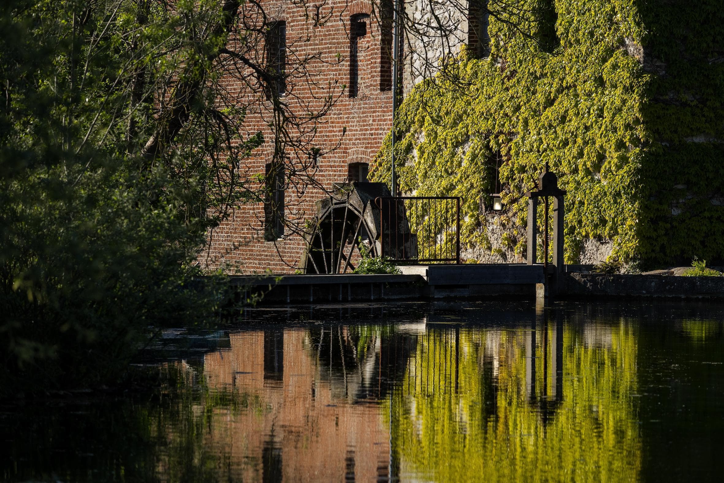 A brick building with a large water wheel is partially covered in green ivy, reflected in the calm water below on a sunny day.