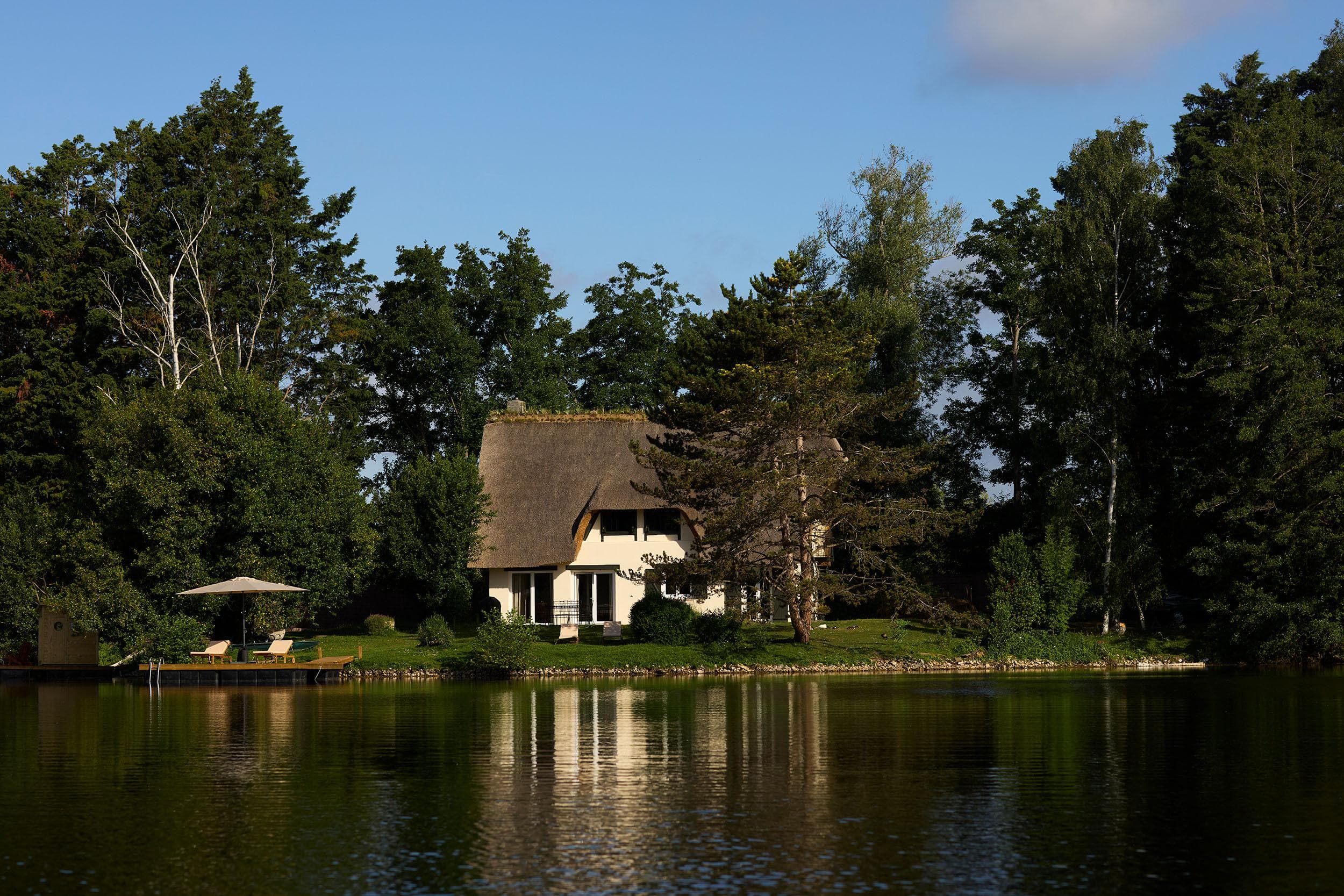 A white cottage with a thatched roof sits surrounded by tall trees on the edge of a calm lake, with its reflection visible in the water and a small dock with an umbrella to the left.