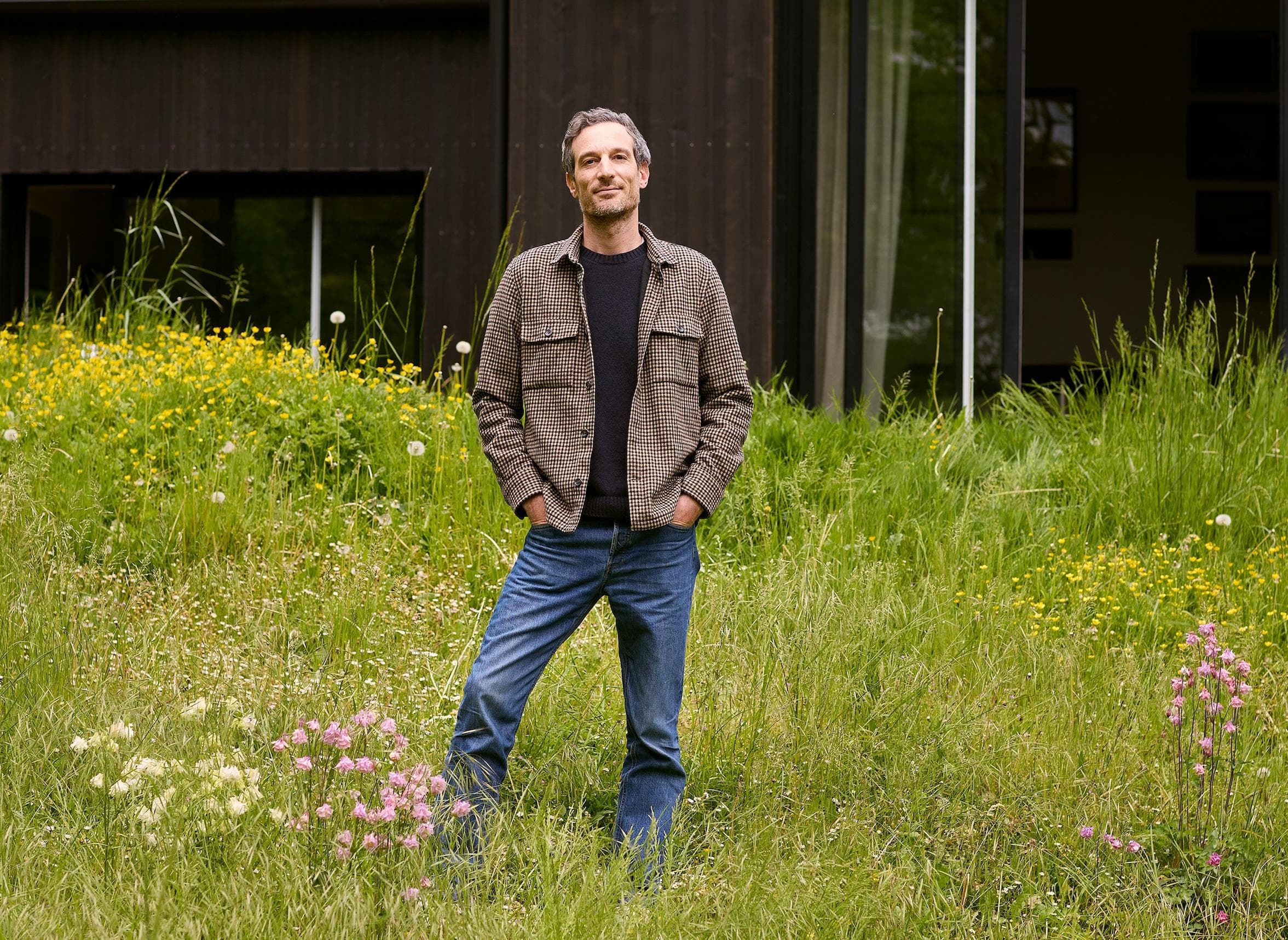 A man stands in tall grass and wildflowers, hands in his pockets, wearing a checked jacket and jeans in front of a modern dark wooden building with large windows.