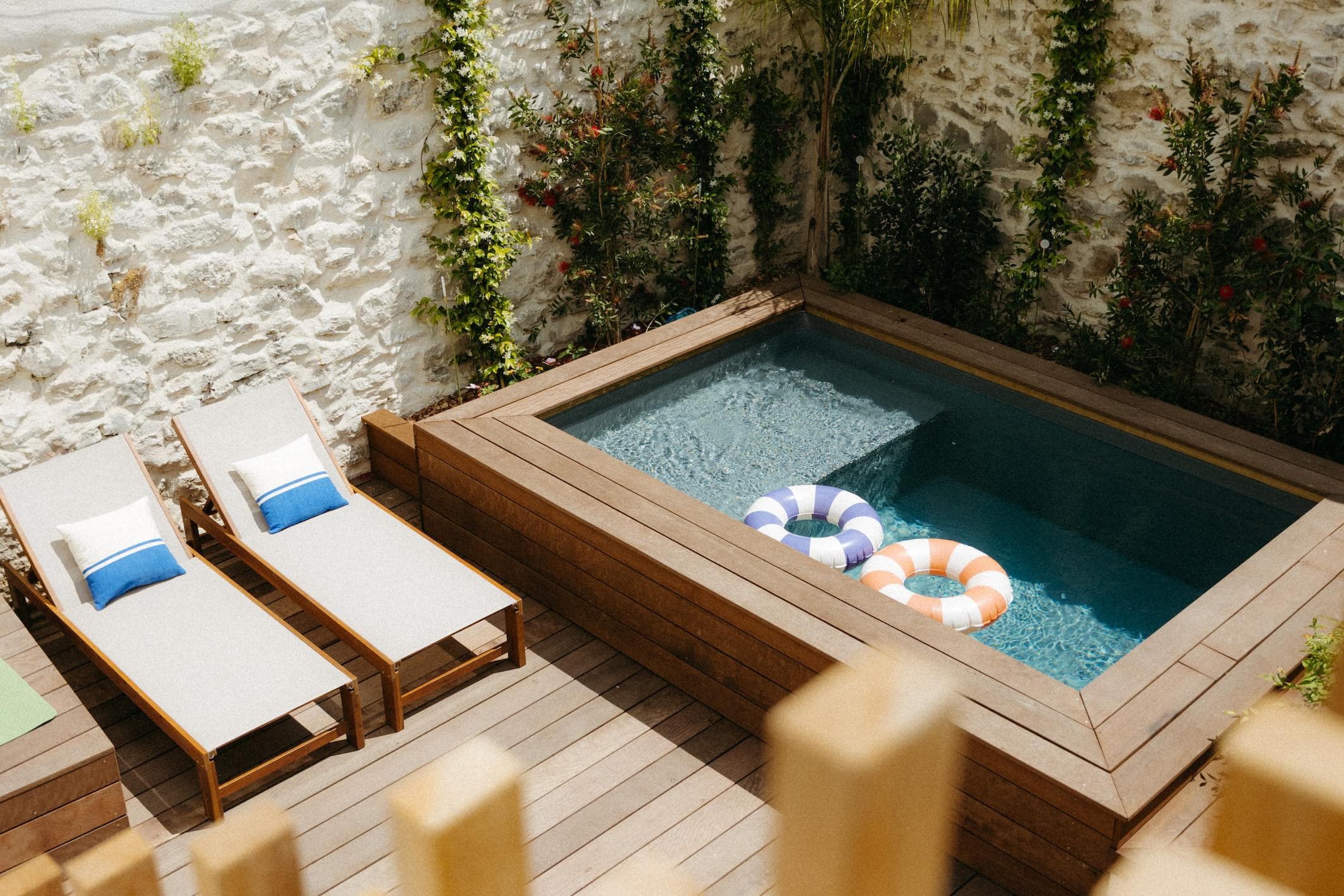 A small rectangular pool with three colorful inflatable rings, surrounded by wooden decking, two lounge chairs with cushions, and stone walls with climbing plants.