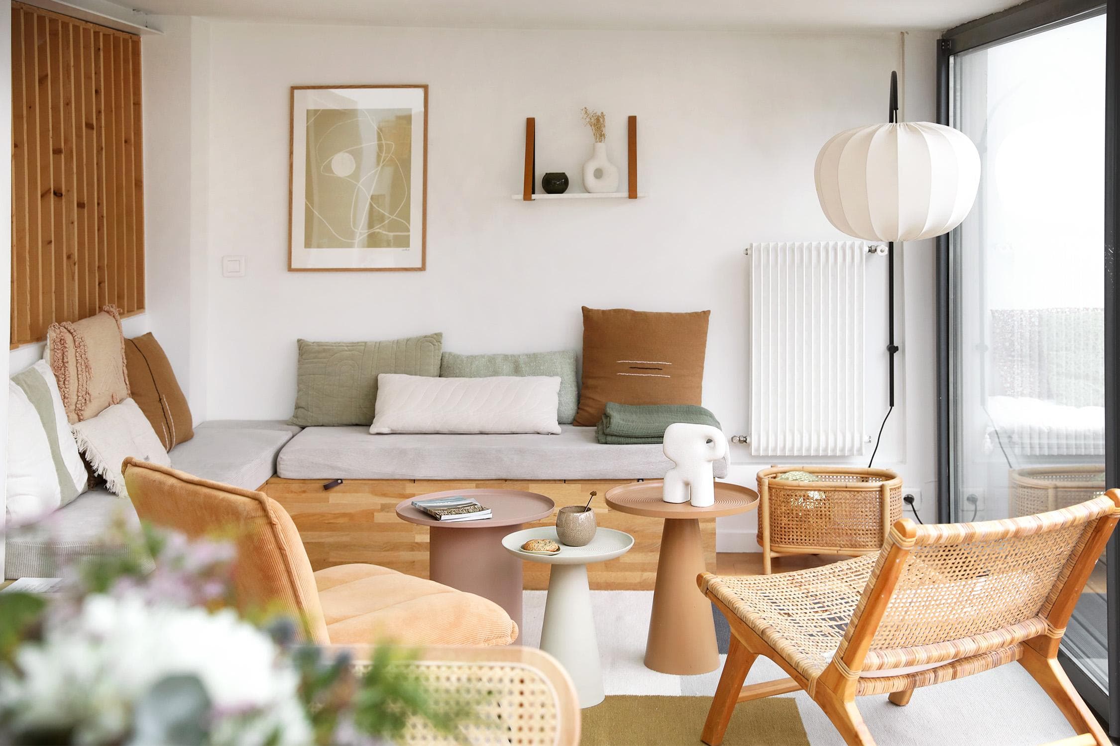 A cozy, modern living room with neutral tones features a built-in cushioned bench, rattan chairs, round coffee tables, abstract art, and a white sculptural lamp by large windows. Decor is minimalist with soft textures.